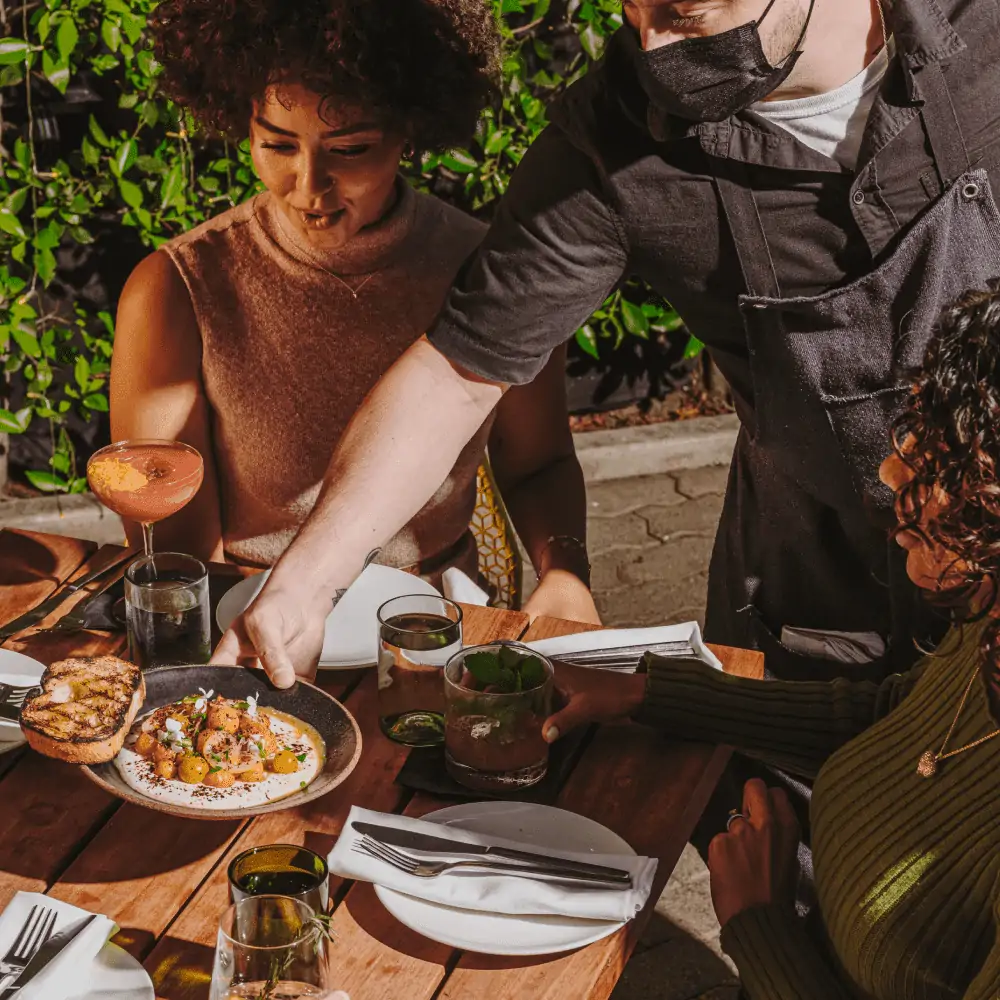 Women sitting at a table being served a beautifully displayed appetizer by a server wearing a mask