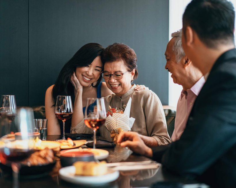 Image depicts a family dining together. They’re sitting in a booth, drinking red wine and sharing small plates. The mother is accepting flowers and she and her daughter smile together and gently embrace. The father and another young man smile as they look on.