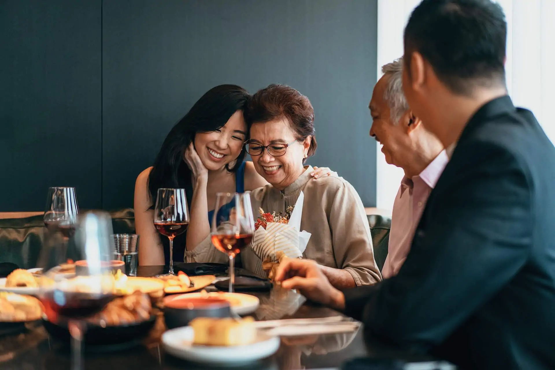 Image depicts a family dining together. They’re sitting in a booth, drinking red wine and sharing small plates. The mother is accepting flowers and she and her daughter smile together and gently embrace. The father and another young man smile as they look on.