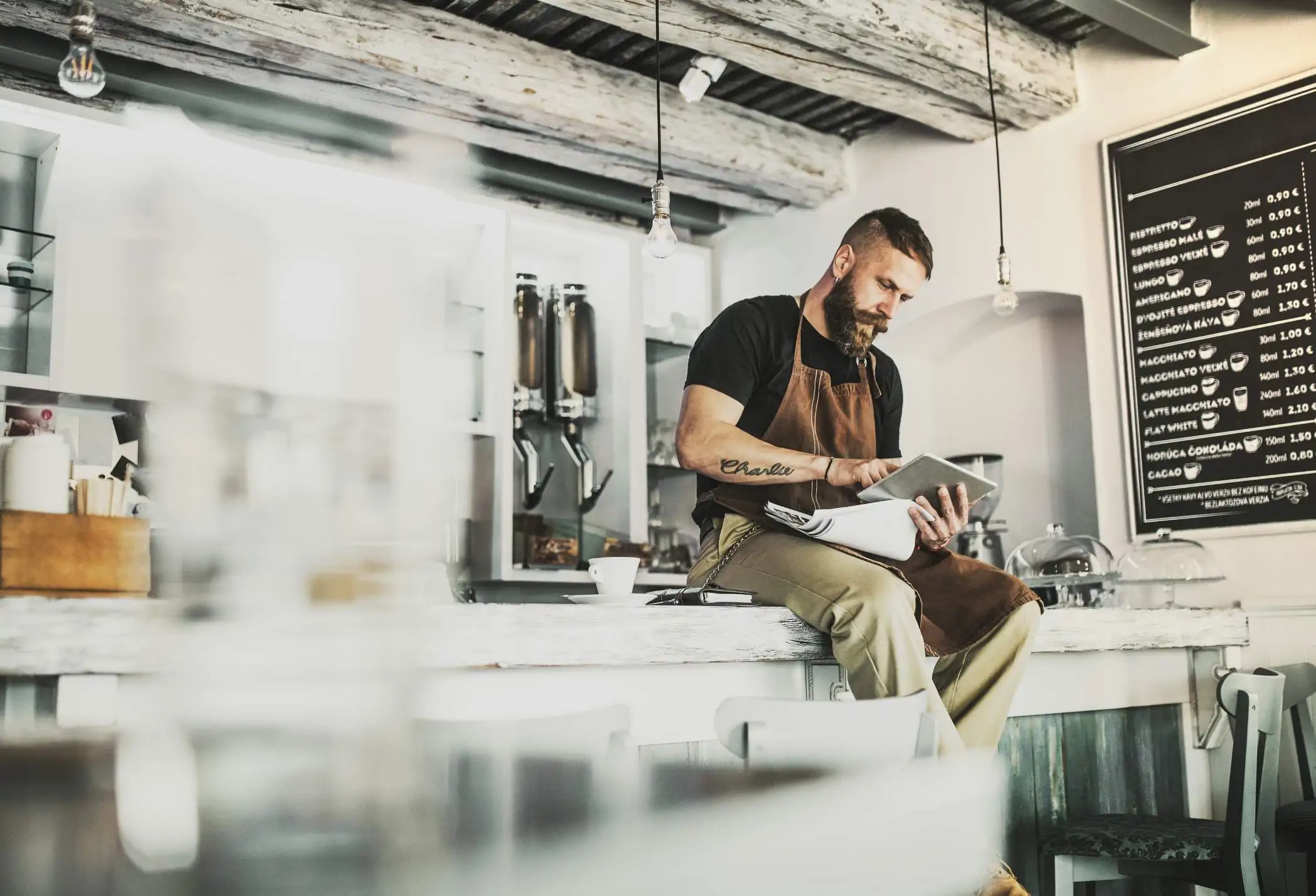 Image depicts a person sitting on a countertop using a tablet. Next to them sits a cup of coffee and a notebook. There is a coffee menu in the back. The establishment is very industrial, with light bulbs hanging from the ceiling.