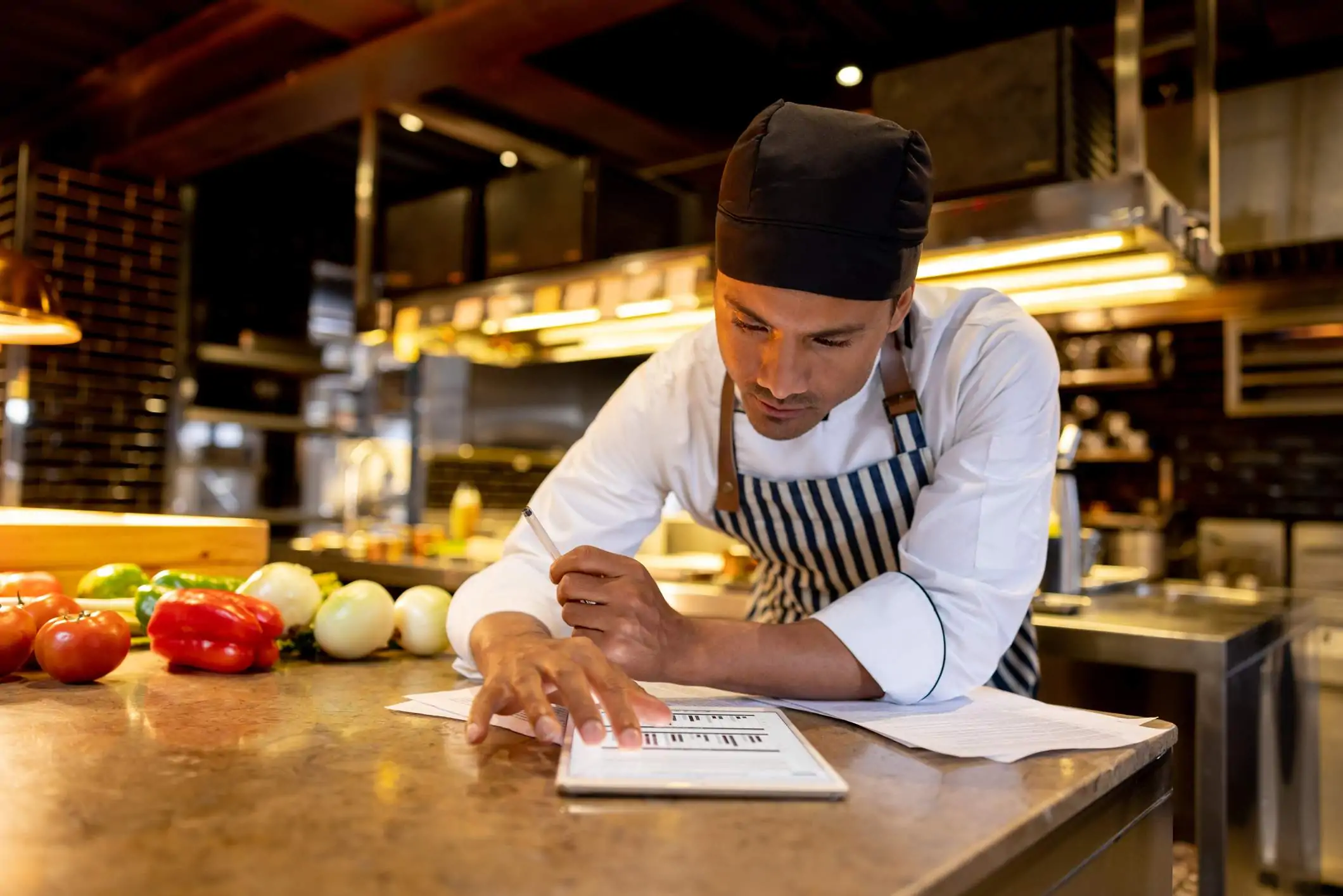 Image depicts a person wearing a black and white striped apron and black cap leaning over a countertop. They are examining graphs on a tablet and there are sheets of paper splayed under them. In the background is an industrial kitchen. There are vegetables sitting on the counter next to them.