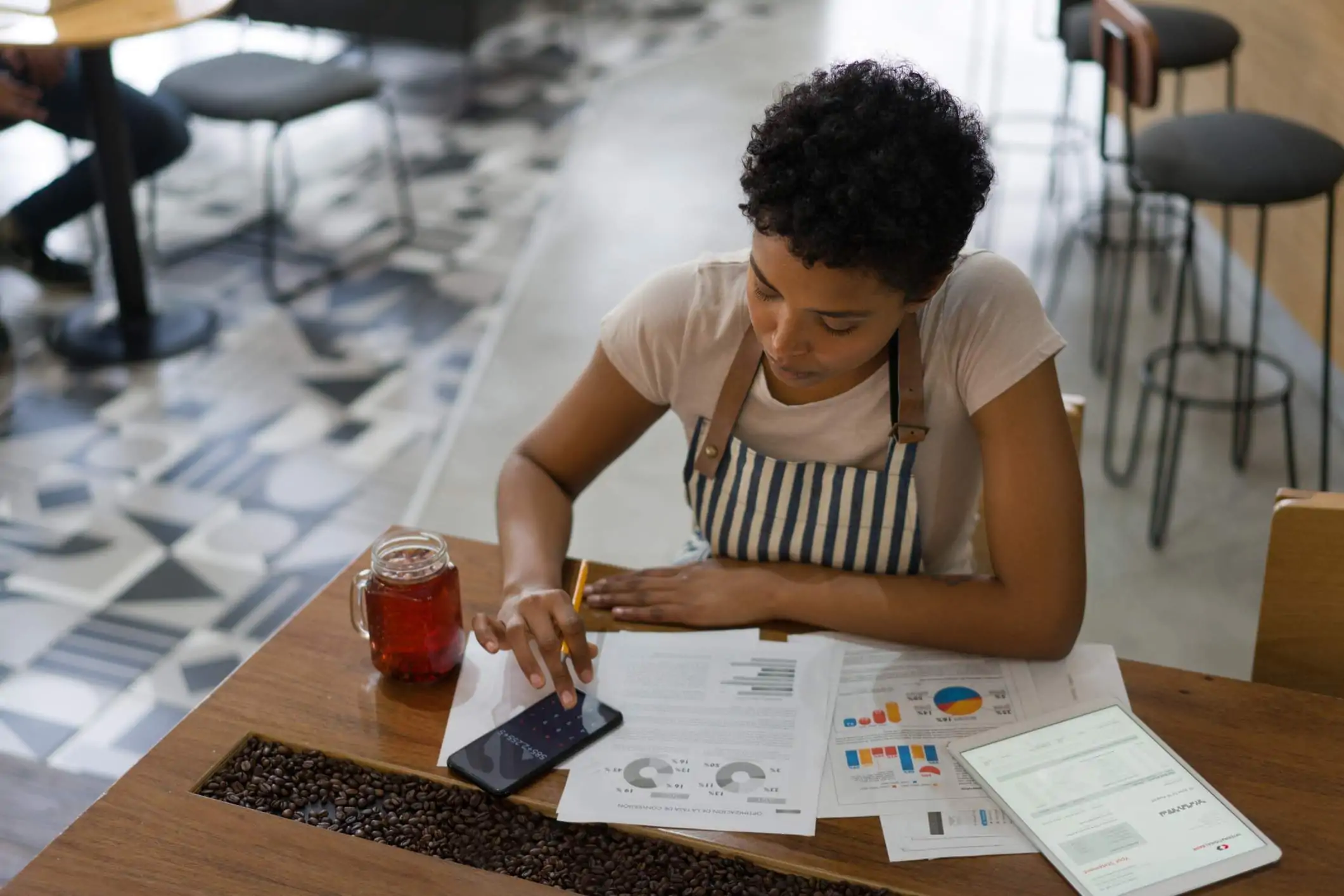 Image depicts a person in a striped apron sitting at a table. There is a mason jar cup with a red drink in it on the table and coffee beans in a slit down the center. The person is calculating something on their phone calculator as sheets of paper and a tablet on the table display different graphs.