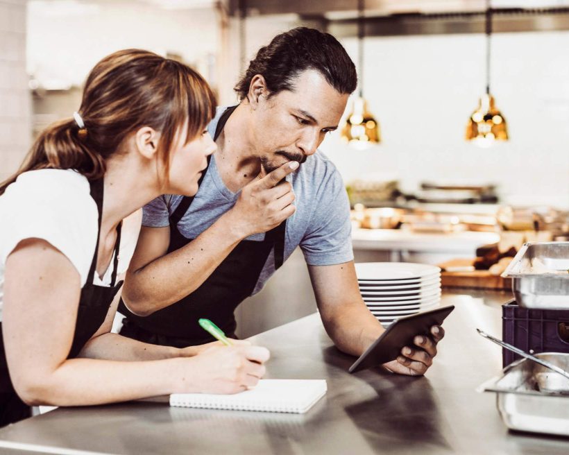 Image depicts two people in a commercial kitchen. They’re both wearing aprons. The one on the left is holding a pen and a pad of paper. The one on the right holds a finger up to their mouth and looks down at a tablet.