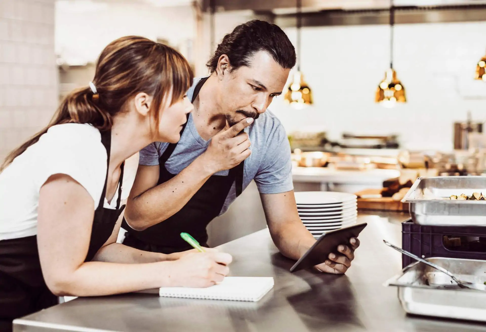 Image depicts two people in a commercial kitchen. They’re both wearing aprons. The one on the left is holding a pen and a pad of paper. The one on the right holds a finger up to their mouth and looks down at a tablet.