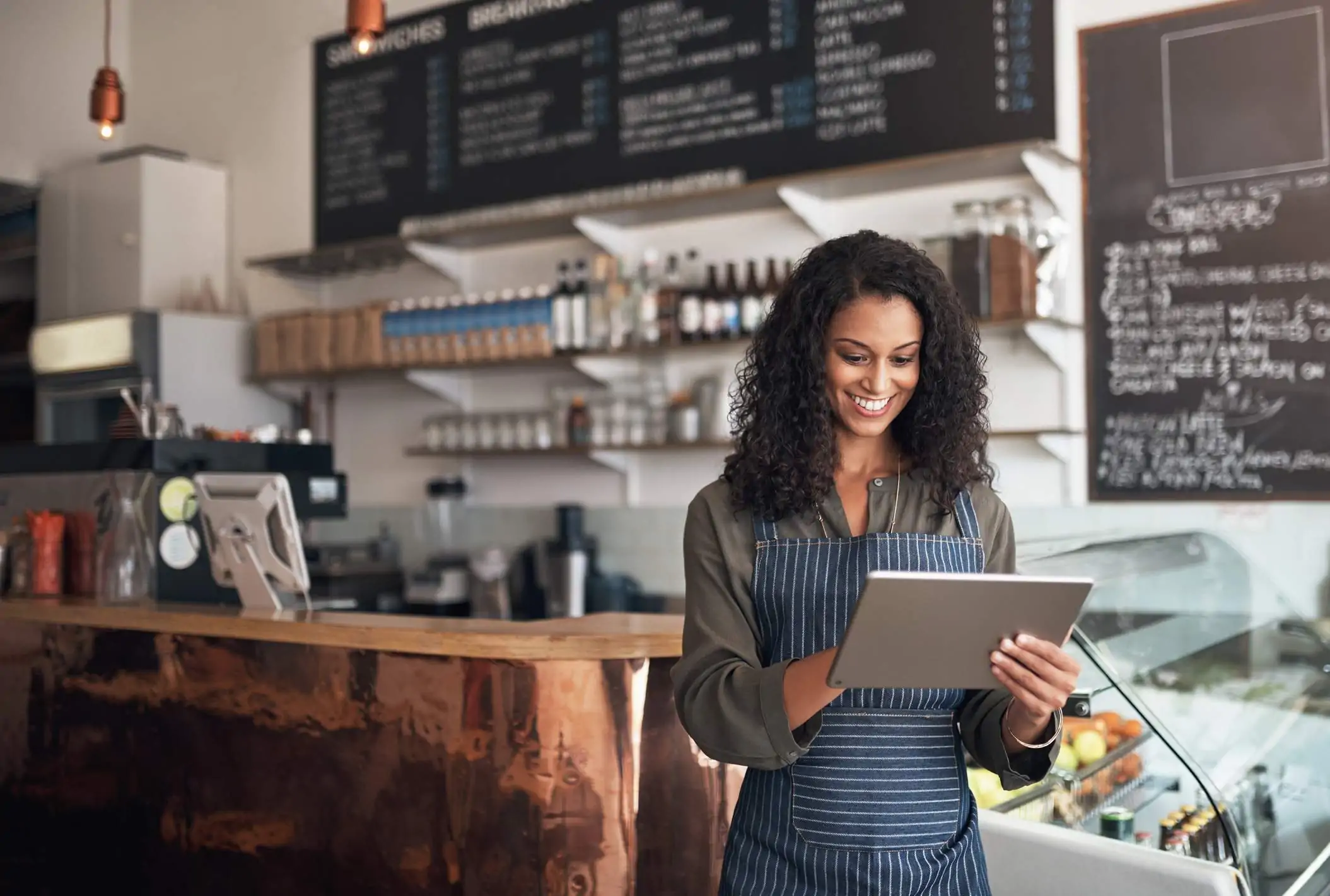 Image depicts a server using an ipad. They are wearing a dark blue apron with white stripes. They are standing inside a fast-casual restaurant.