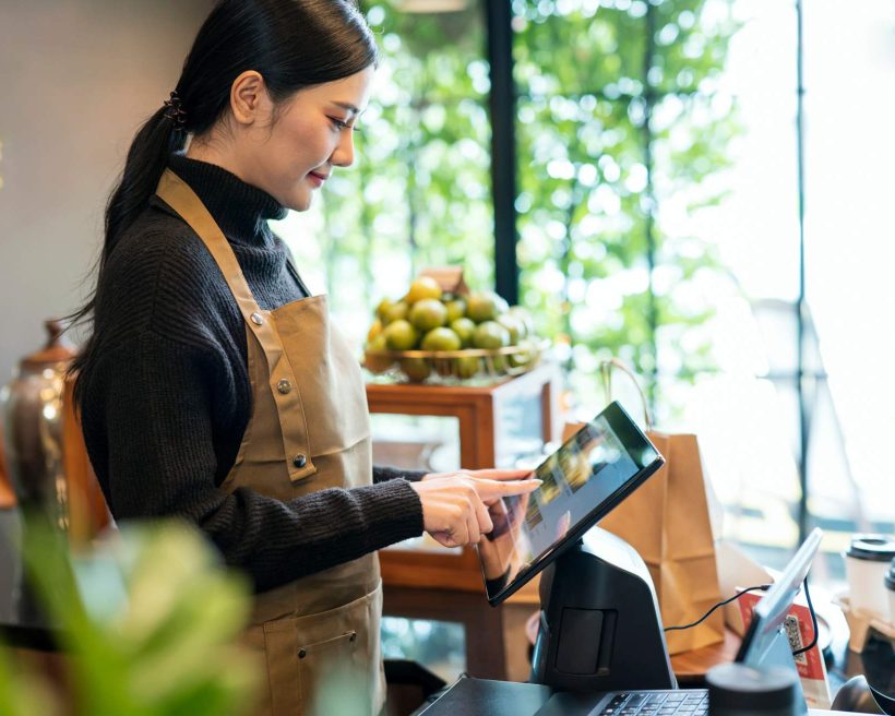 Image depicts a person wearing a black turtleneck and a brown apron using a mounted tablet in a restaurant.
