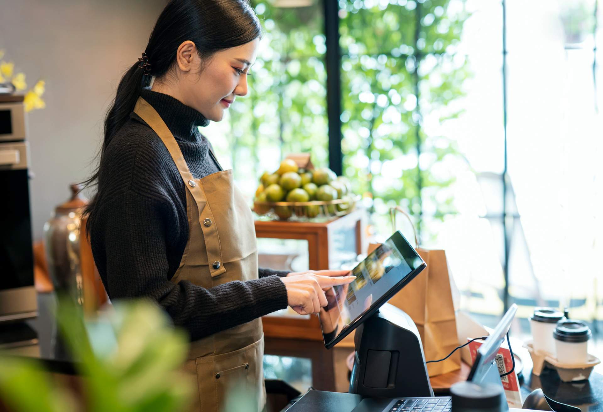 Image depicts a person wearing a black turtleneck and a brown apron using a mounted tablet in a restaurant.
