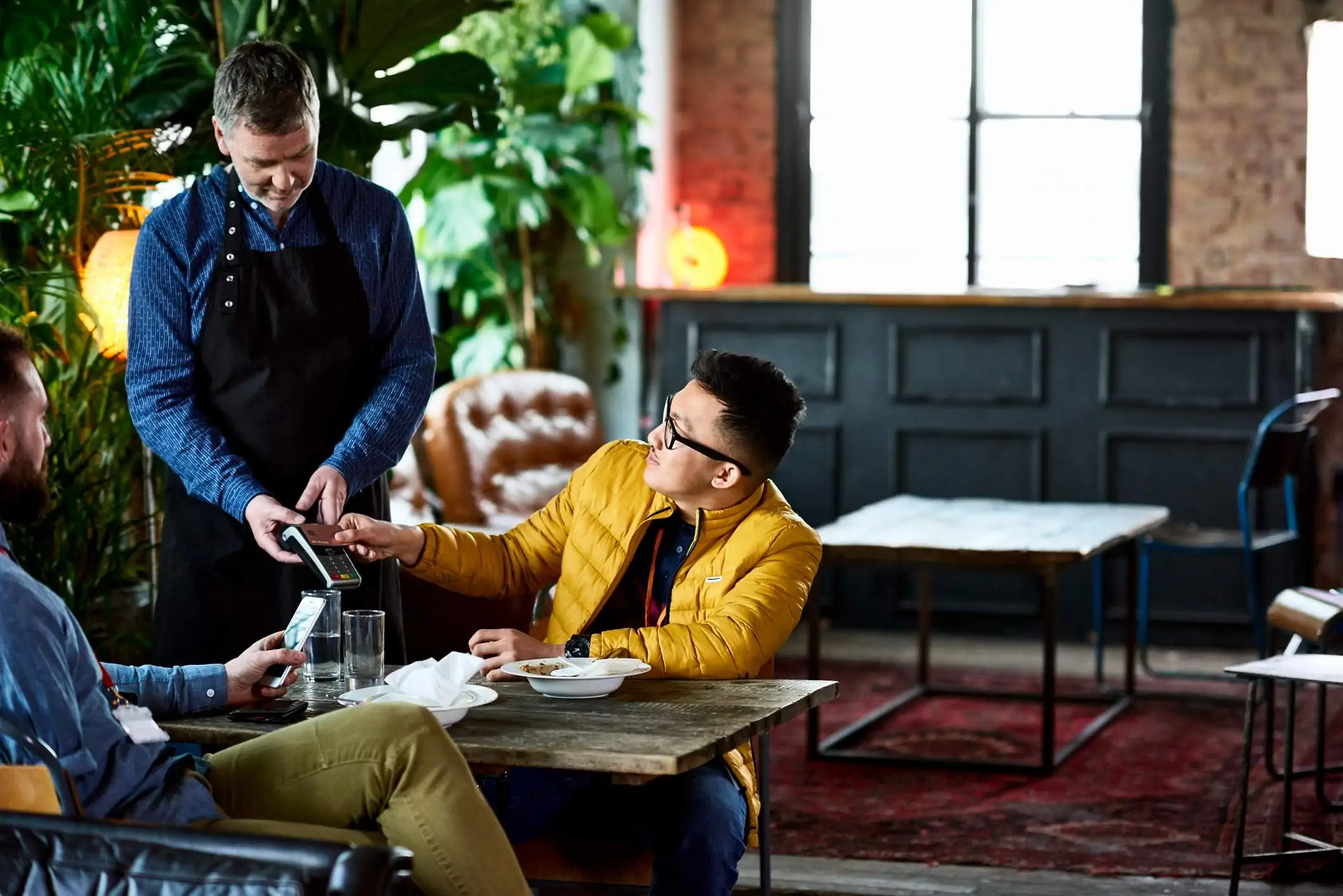 Image depicts a person in a yellow puffer jacket and black rimmed glasses utilizing a point-of-sale (POS) system to pay for a meal at a restaurant. A server wearing a blue button down and black apron is holding it out to the guest. The other diner at the table is looking at their phone.