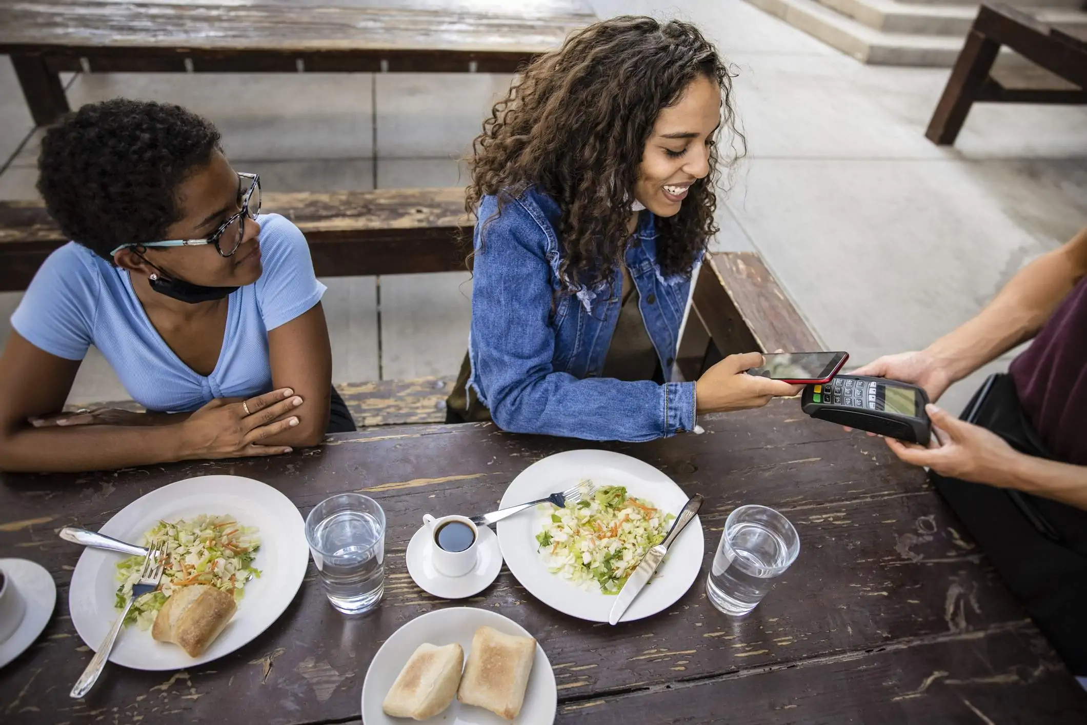 Image depicts two guests sitting at a table outside of a restaurant. They are eating salads and bread. The diner on the right is paying for the meal using their phone and a point-of-sale (POS) system. The server, standing out of frame, is holding the device out to the guest.