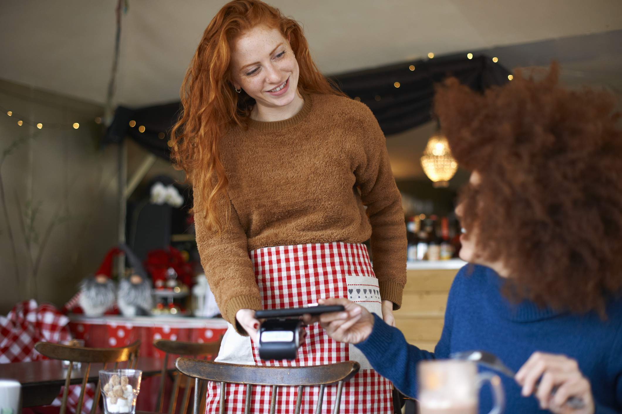 Image depicts a server wearing an orange sweater and a red plaid apron holding out a point-of-sale (POS) system to a guest. The diner is wearing a blue sweater and is tapping their phone to the device to pay for the meal. 