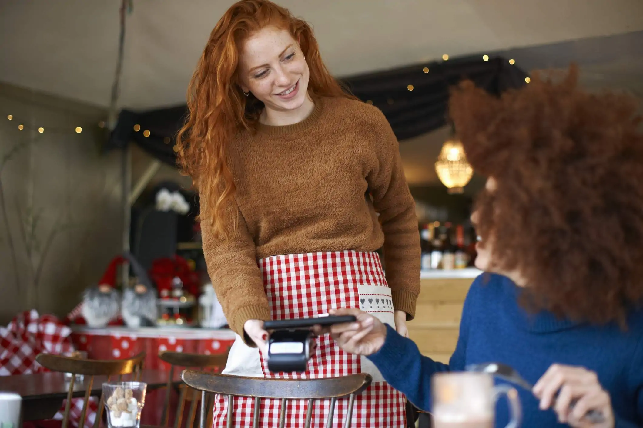 Image depicts a server wearing an orange sweater and a red plaid apron holding out a point-of-sale (POS) system to a guest. The diner is wearing a blue sweater and is tapping their phone to the device to pay for the meal.