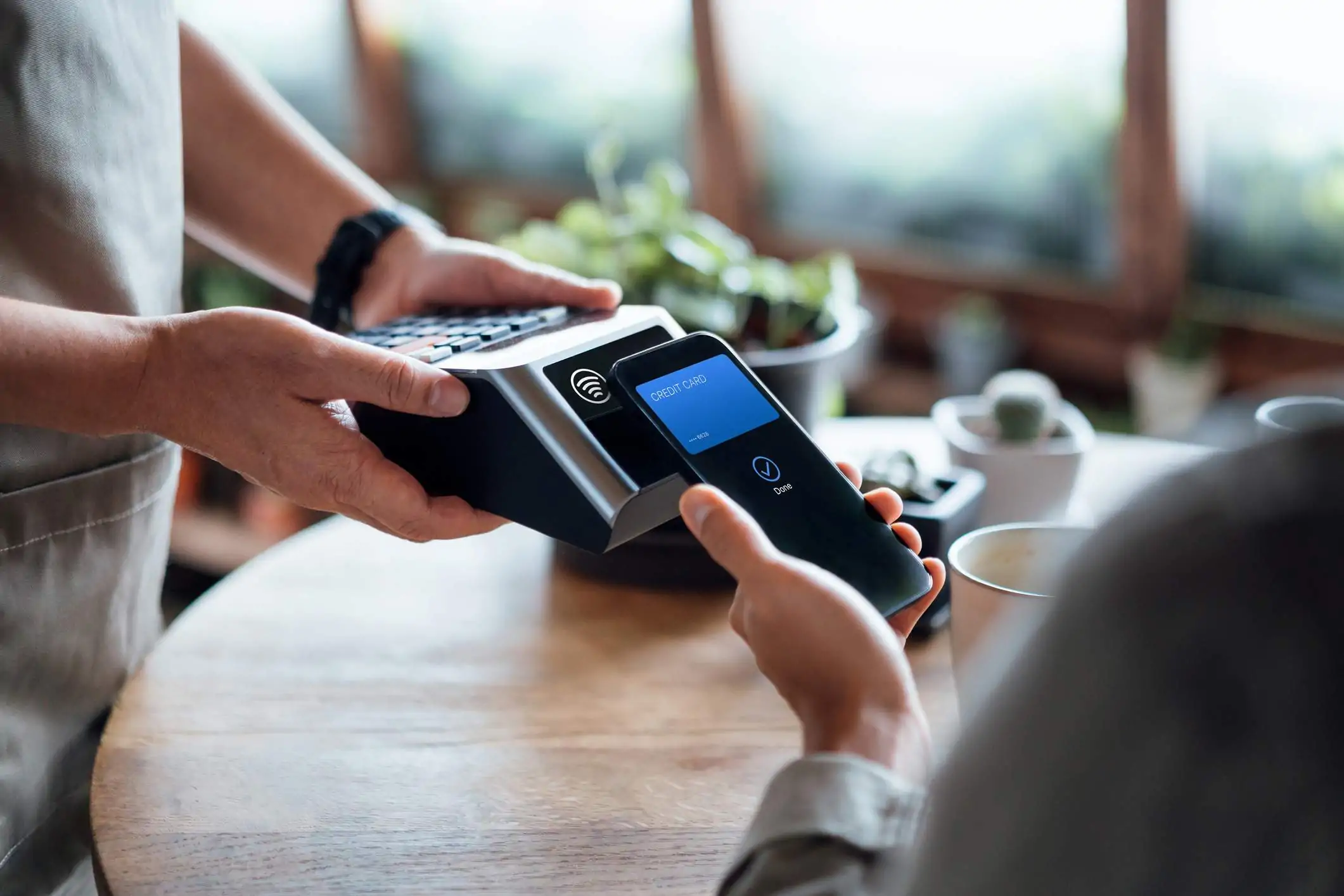 Image depicts two people standing out of frame. One looks to be a server and is holding out a point-of-sale (POS) system to a guest. The guest is holding their phone out to pay for their meal.