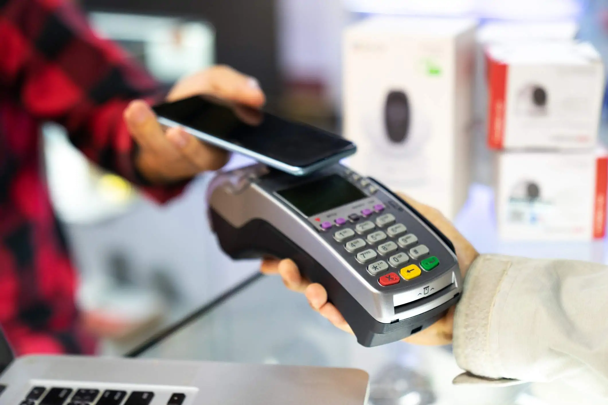 Image depicts two people standing out of frame. One looks to be a server and is holding out a point-of-sale (POS) system to a guest. The guest, wearing a red plaid shirt, is holding their phone out to pay for their meal.