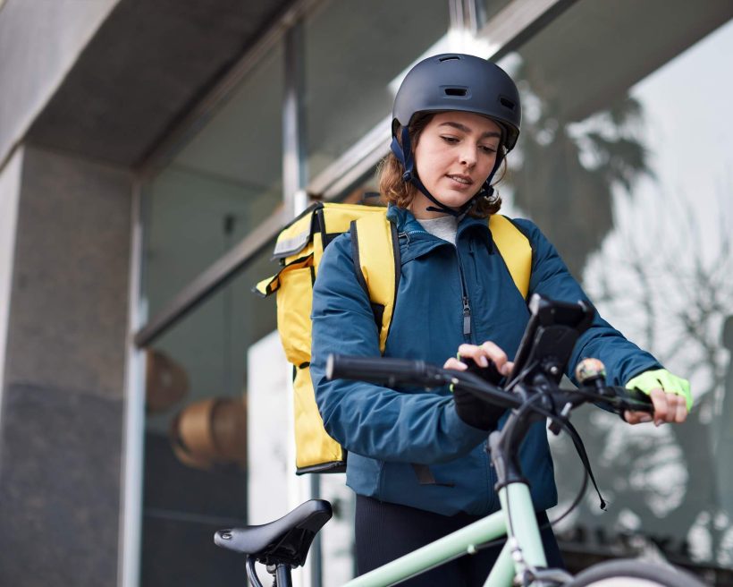 Image depicts a delivery driver standing next to their bike. They are using the mobile phone that is mounted on the front of the bike. They are wearing a black helmet and a yellow delivery box is strapped to their back.