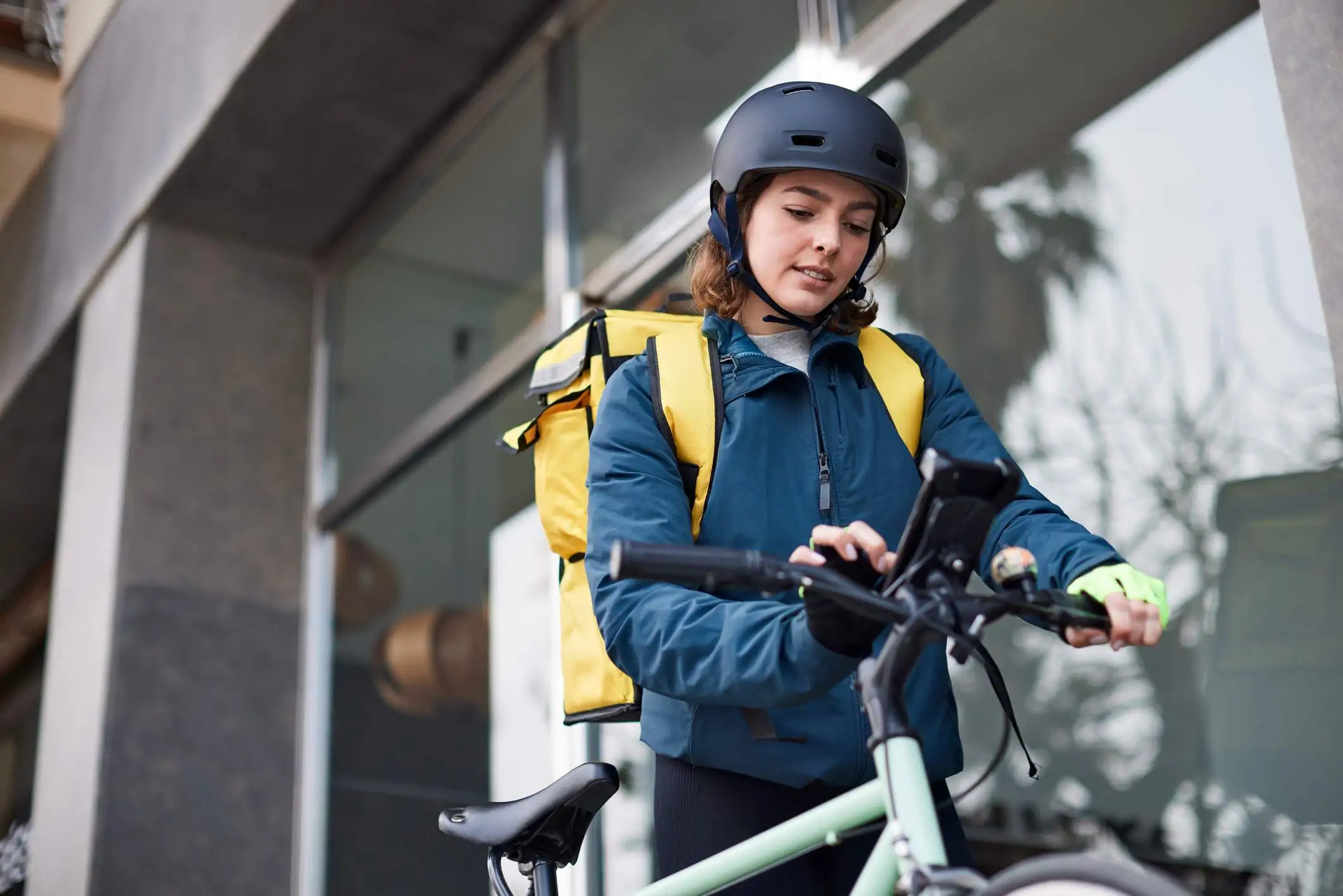 Image depicts a delivery driver standing next to their bike. They are using the mobile phone that is mounted on the front of the bike. They are wearing a black helmet and a yellow delivery box is strapped to their back.