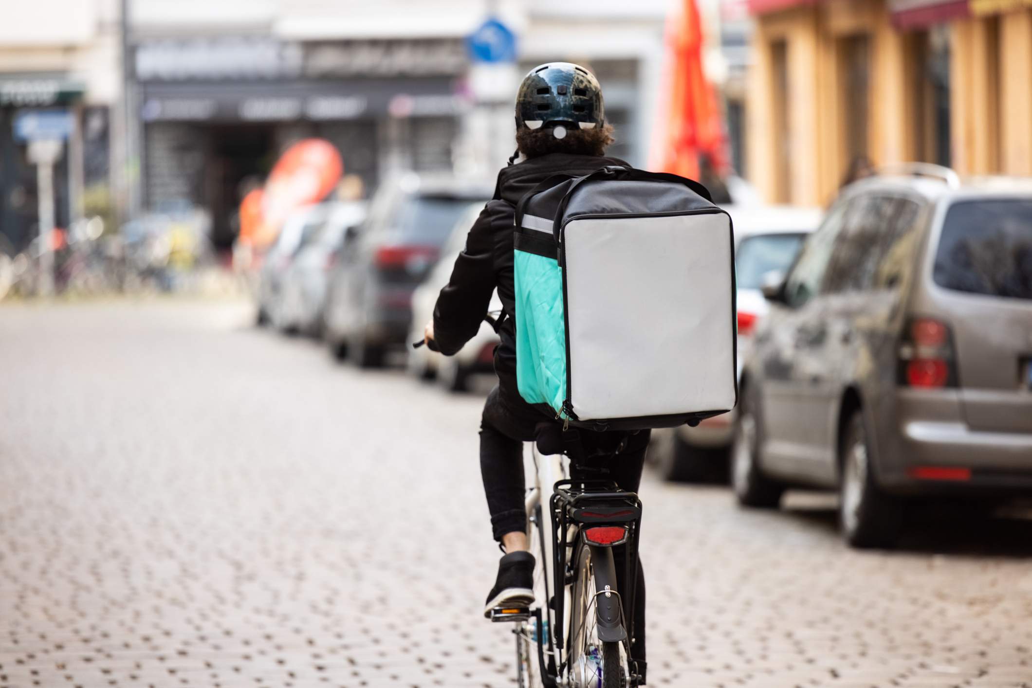 Image depicts a delivery driver with a teal cooler strapped to their back, They are biking down a cobblestone street. 