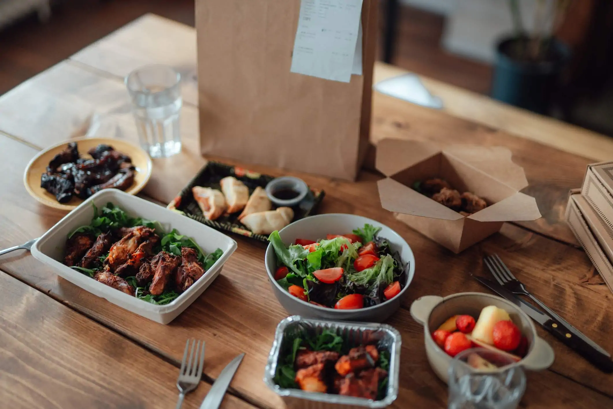 Image depicts plates of takeout sitting on a table. There are some dishes of meat and some vegetables. There is a paper bag sitting in the back.