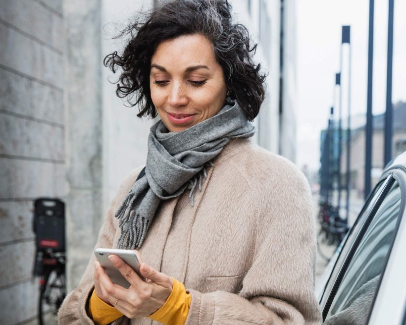 Image depicts a person holding a phone outside. They are wearing a coat and scarf and stand next to a car. They are looking down at the piece of technology in their hands.
