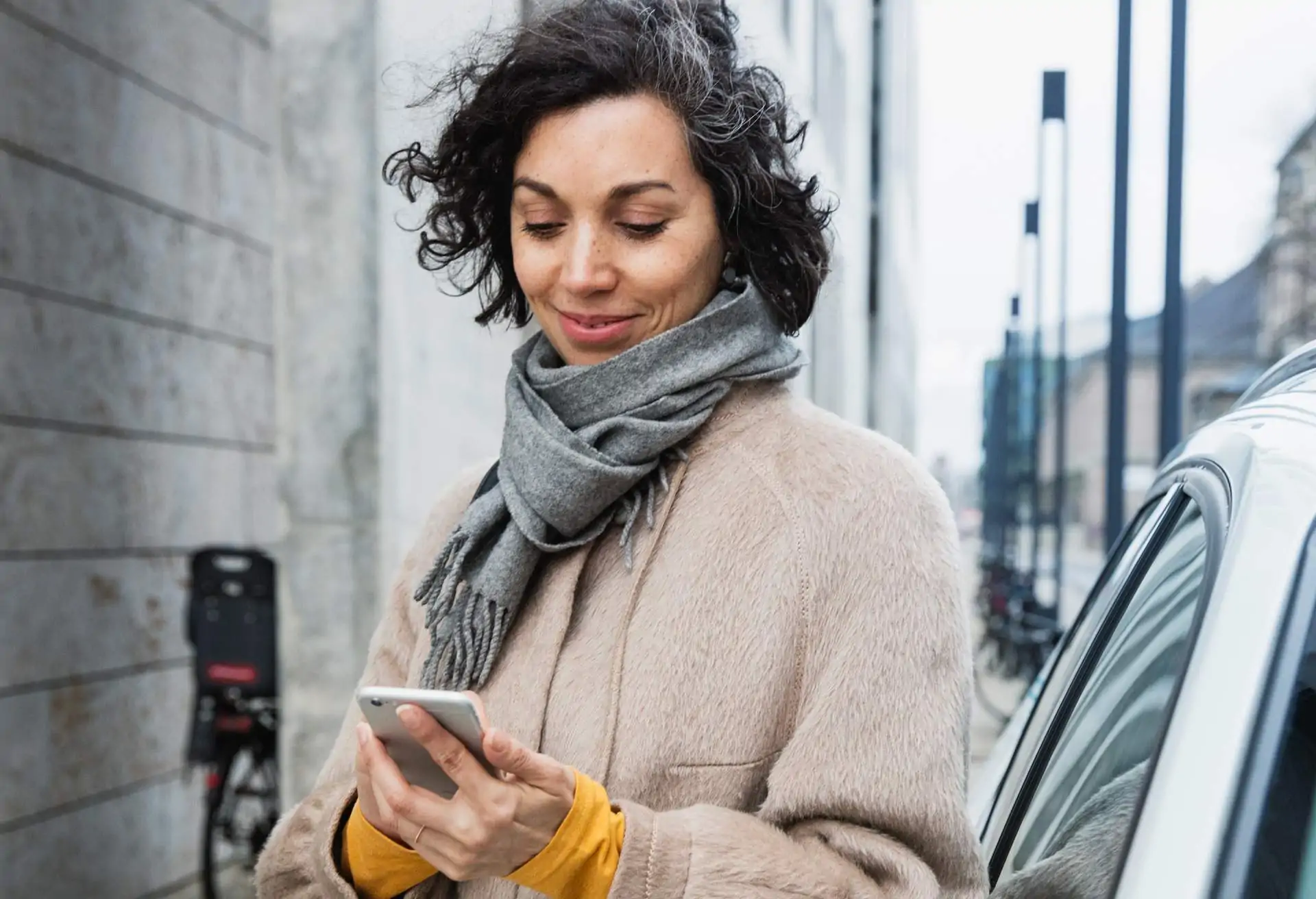 Image depicts a person holding a phone outside. They are wearing a coat and scarf and stand next to a car. They are looking down at the piece of technology in their hands.