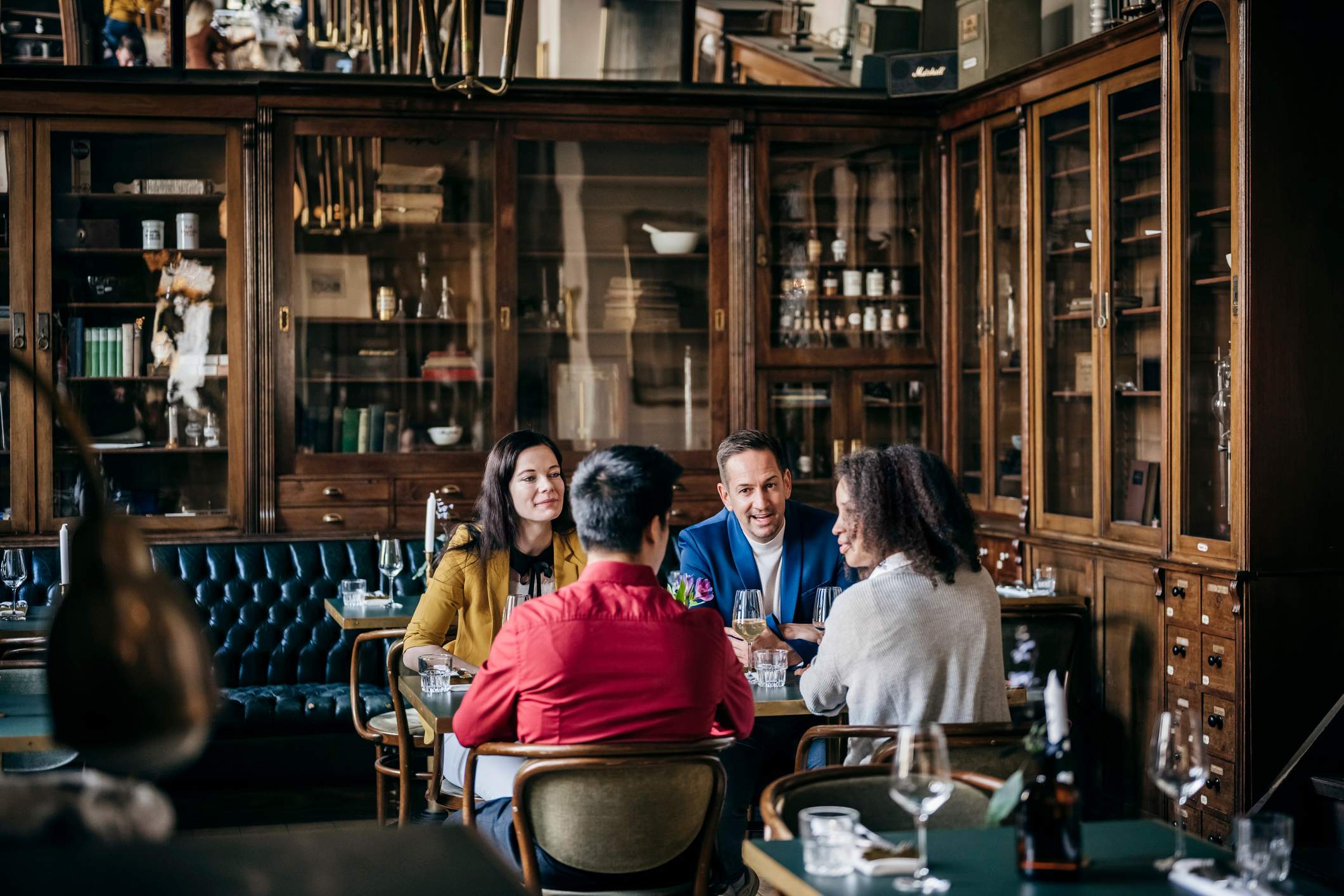 Image depicts four diners sitting together at a table. The restaurant has a vintage office feel, with leather booths and bookcases built into the walls. The diners are all wearing different colors and are chatting amongst themselves. 
