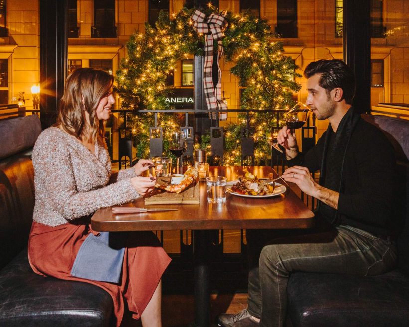 Image depicts two people sharing dinner during the holiday season. They are both drinking wine and are seated in a restaurant booth next to a large window. There is a large wreath with a plaid bow hanging behind them.