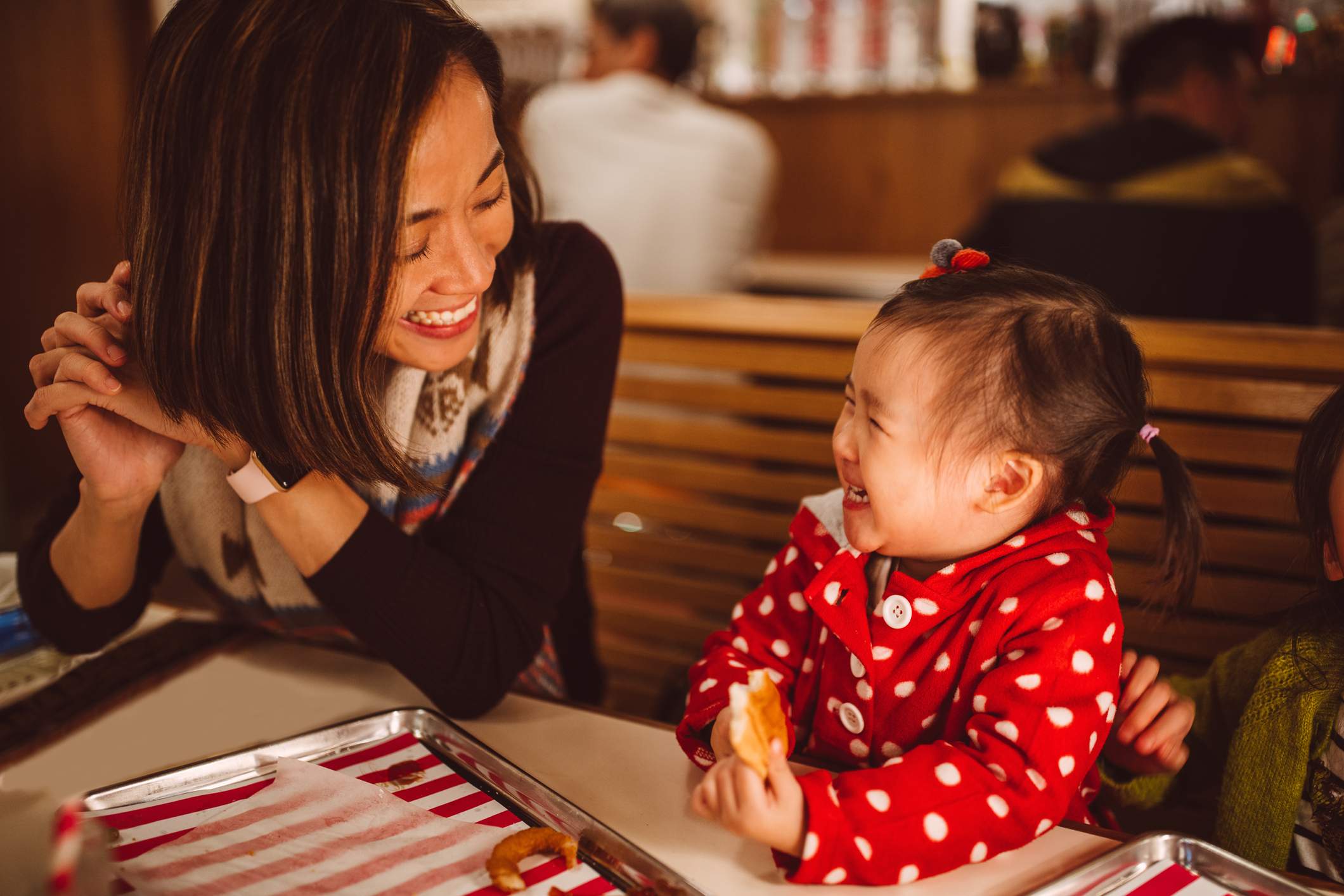Image depicts a small child in a red polkadot jacket eating some bread and smiling at their parent. Their parent sits in the booth next to them and is smiling back. 
