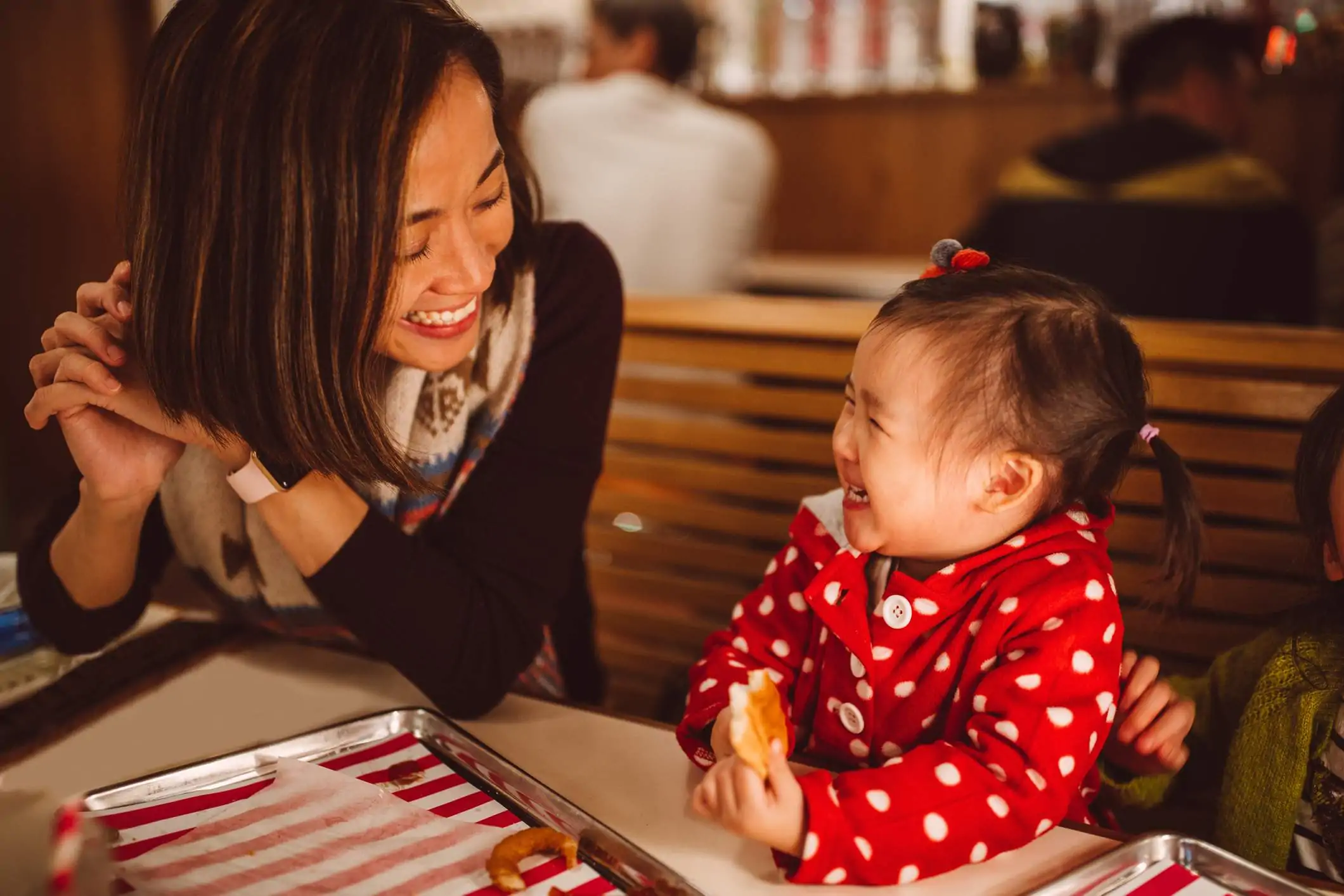 Image depicts a small child in a red polkadot jacket eating some bread and smiling at their parent. Their parent sits in the booth next to them and is smiling back.