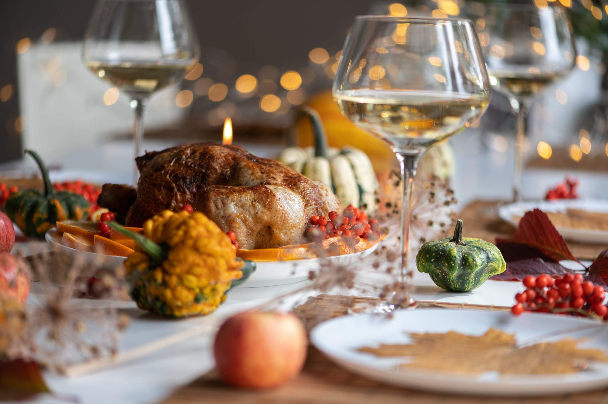 Image depicts a table set for the holidays. There is a cooked bird in the middle of the table and gords sprinkled around as decorations. There is white wine in the glasses. 