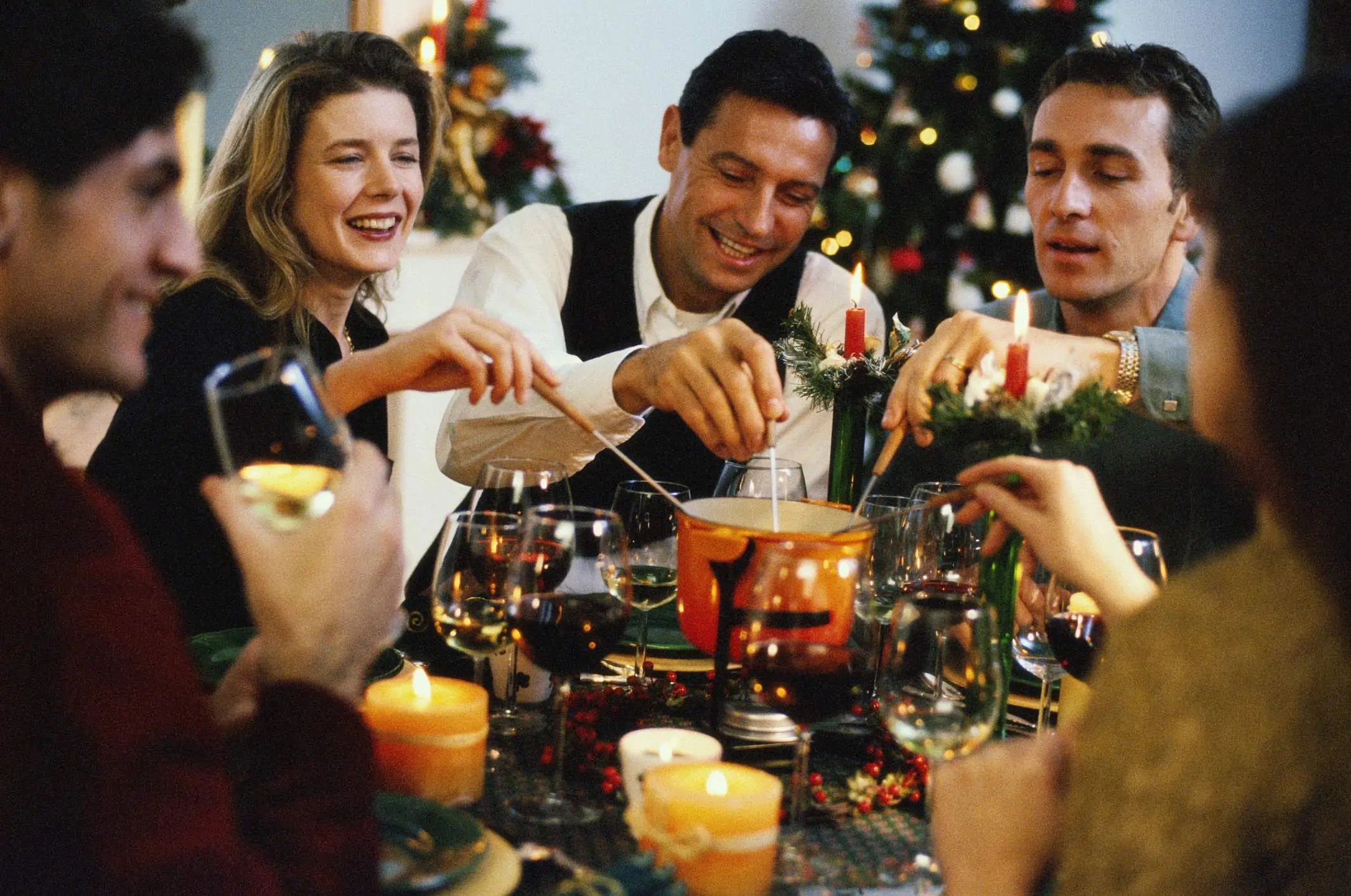 Image depicts six people sitting at a dining table. They are drinking a mix of wines and four of the people are eating the fondue in the middle of the table. There are lit candles strewn throughout.