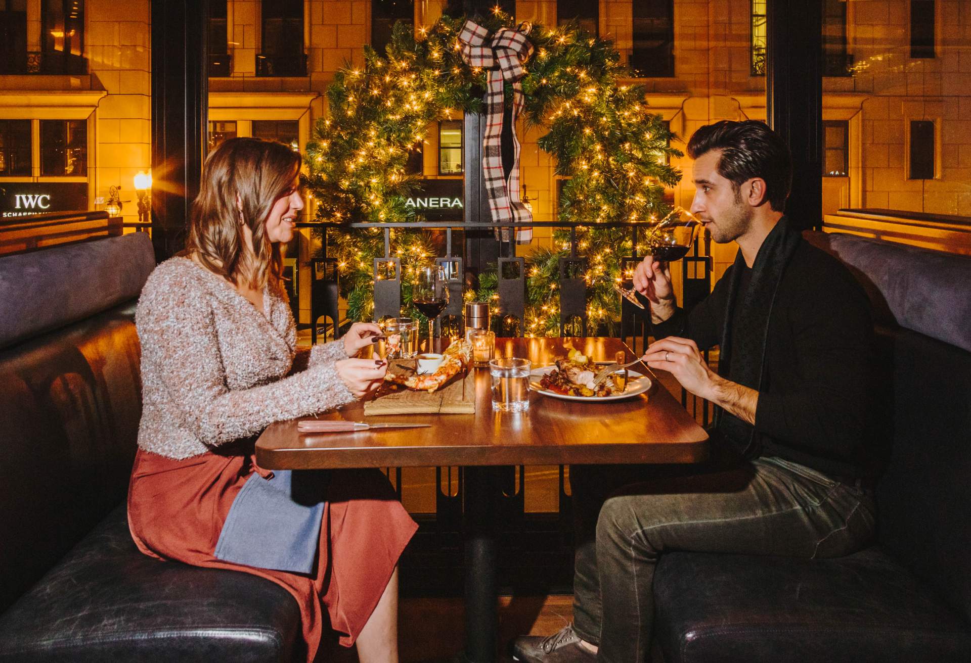 Image depicts two people sharing dinner during the holiday season. They are both drinking wine and are seated in a restaurant booth next to a large window. There is a large wreath with a plaid bow hanging behind them.