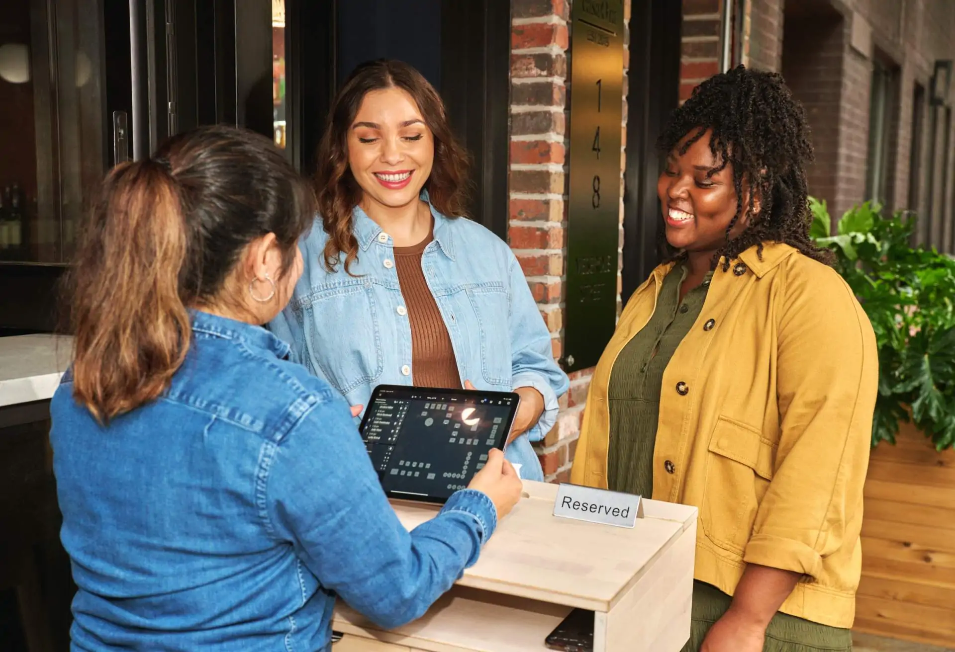 Image depicts two people approaching the host stand. They are both smiling as the host checks a tablet for their reservation.