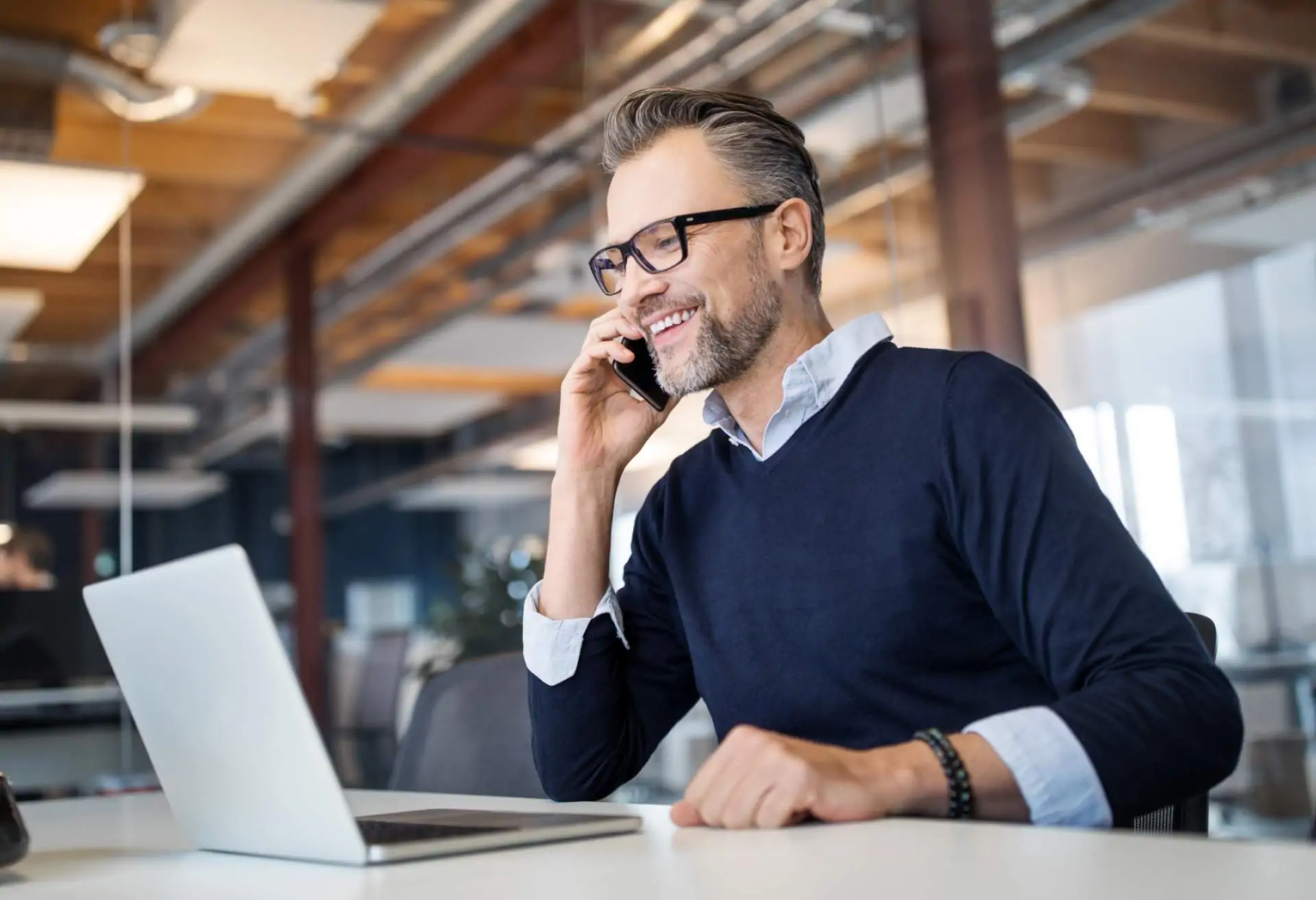 Image depicts a person wearing black rimmed glasses speaking on a mobile phone. There is a laptop on the table in front of them.