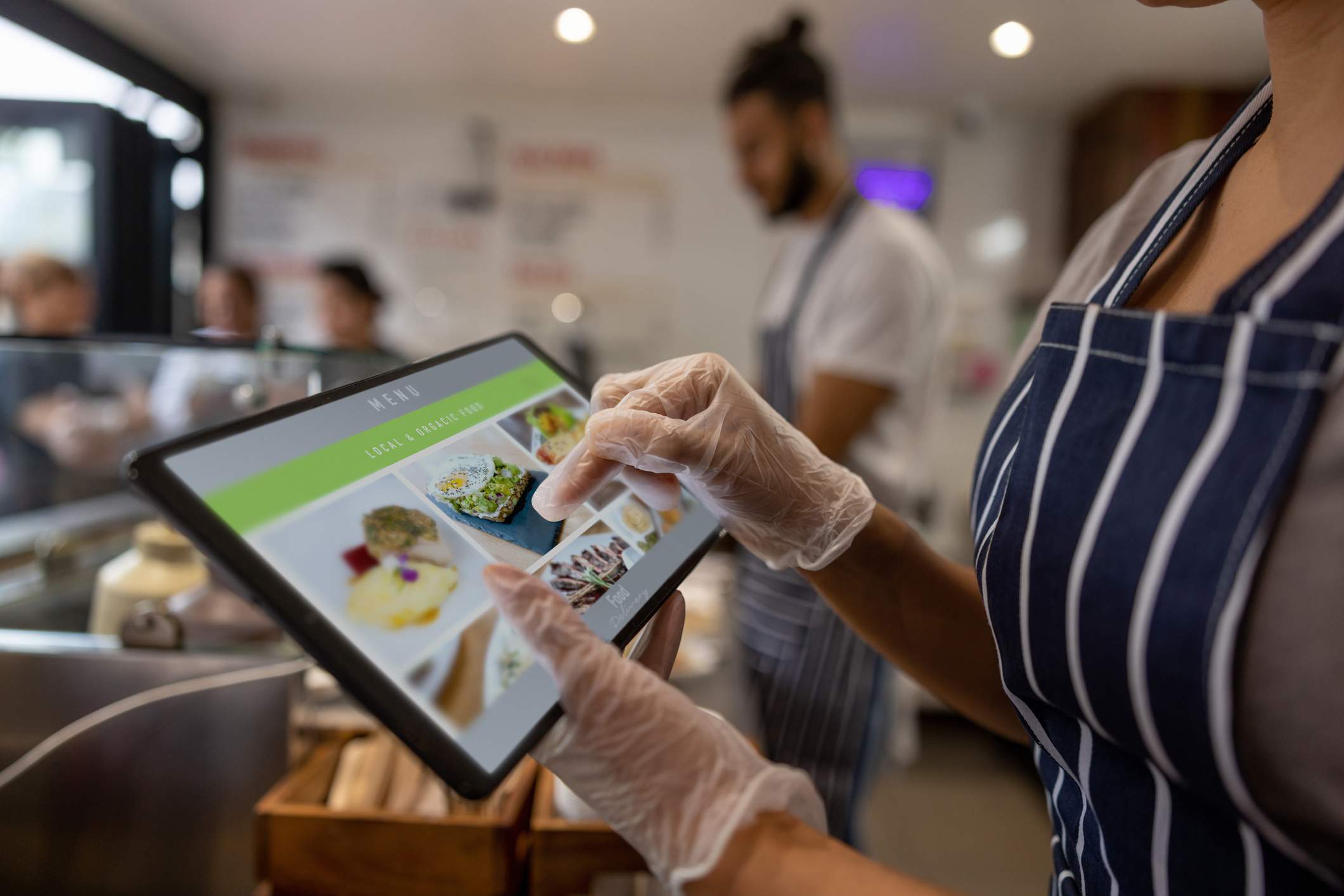 Image depicts a server wearing a blue and white striped apron and plastic gloves using a tablet. The tablet displays different menu options.