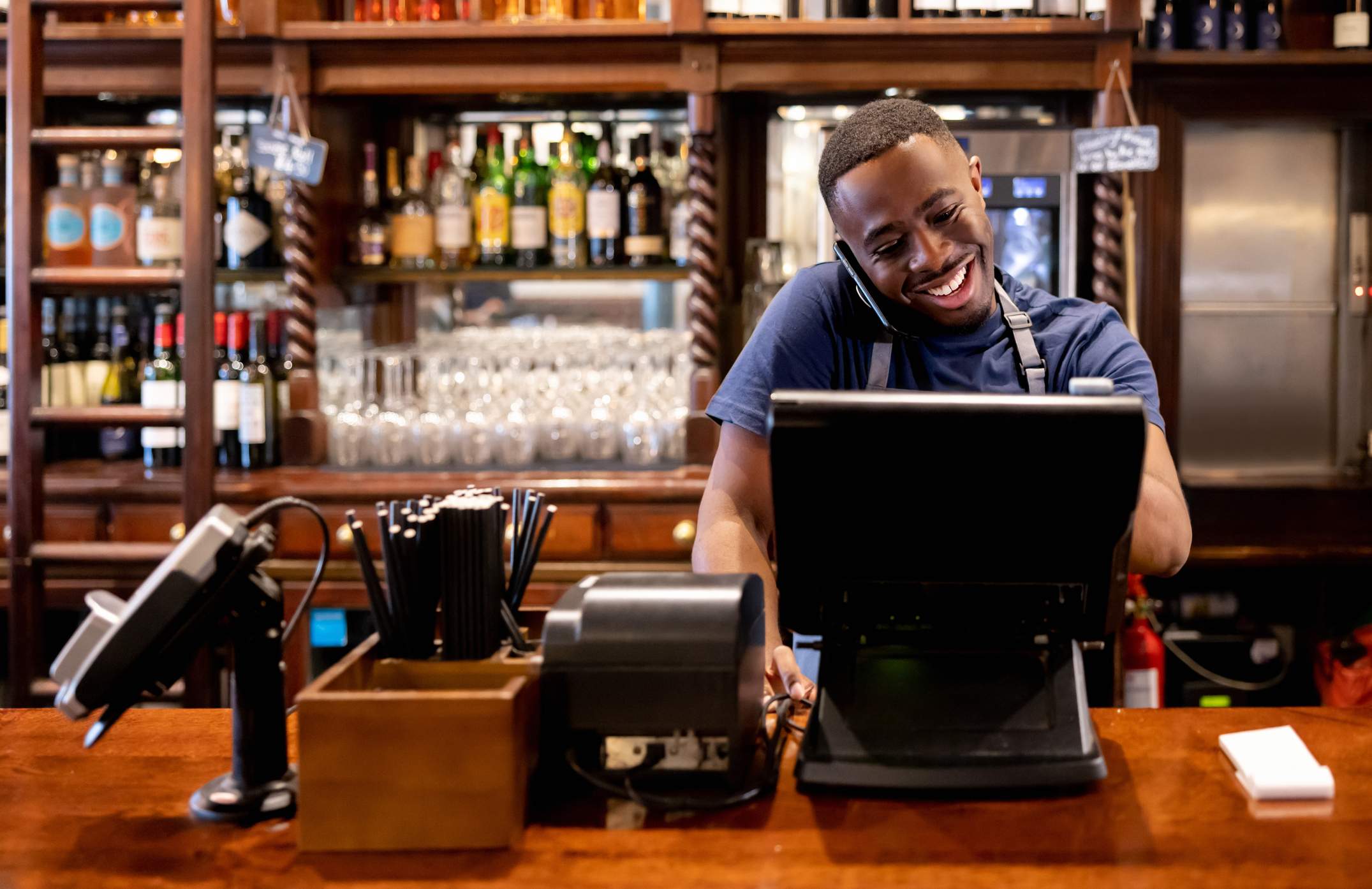 Image depicts a server wearing a blue t-shirt and black apron speaking to someone on the phone while they use a computer in front of them. There is a bar set up behind them. 
