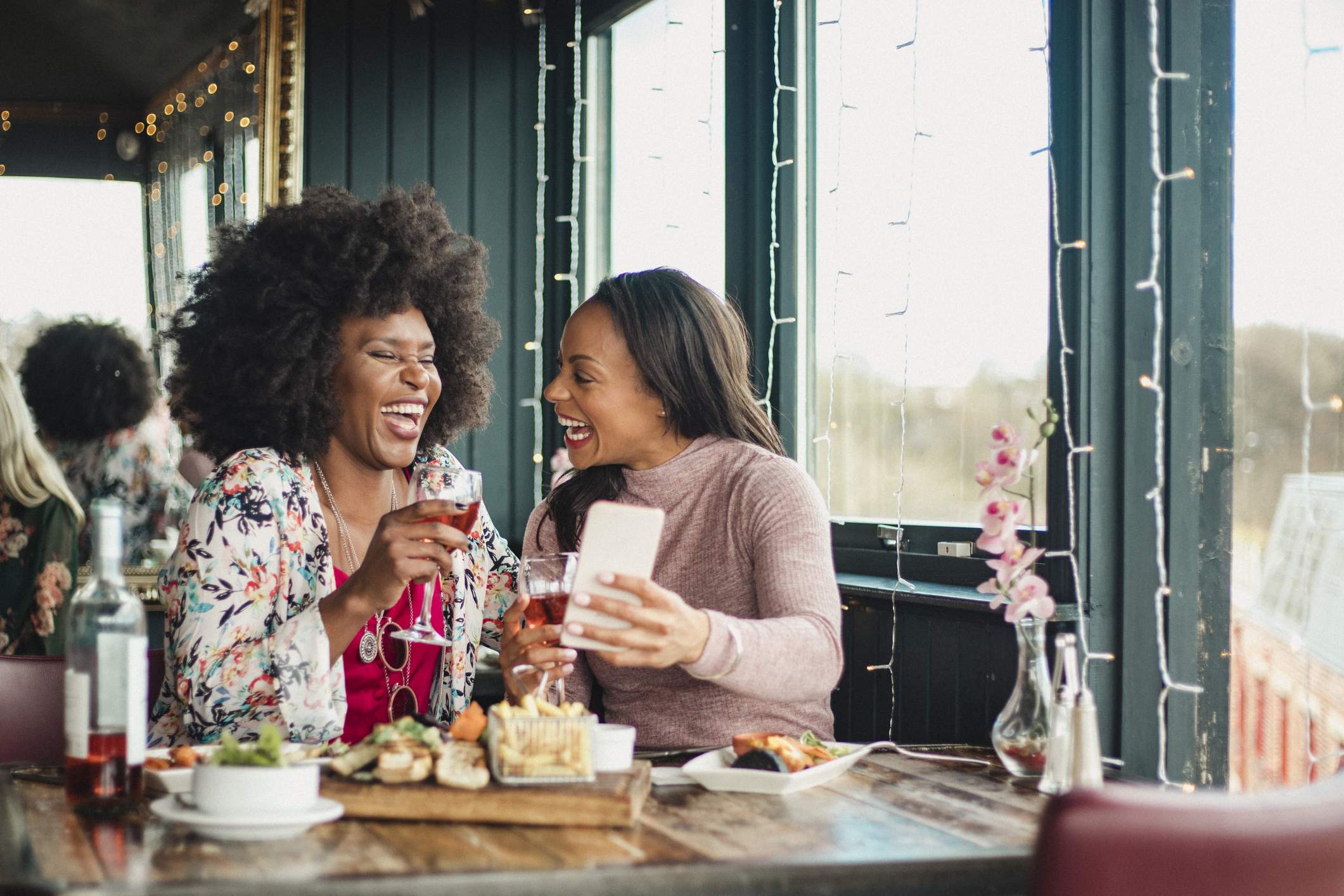 Image depicts two diners seated at a restaurant. They are sharing some plates and drinking wine. They are both laughing as one of them holds up their phone. 