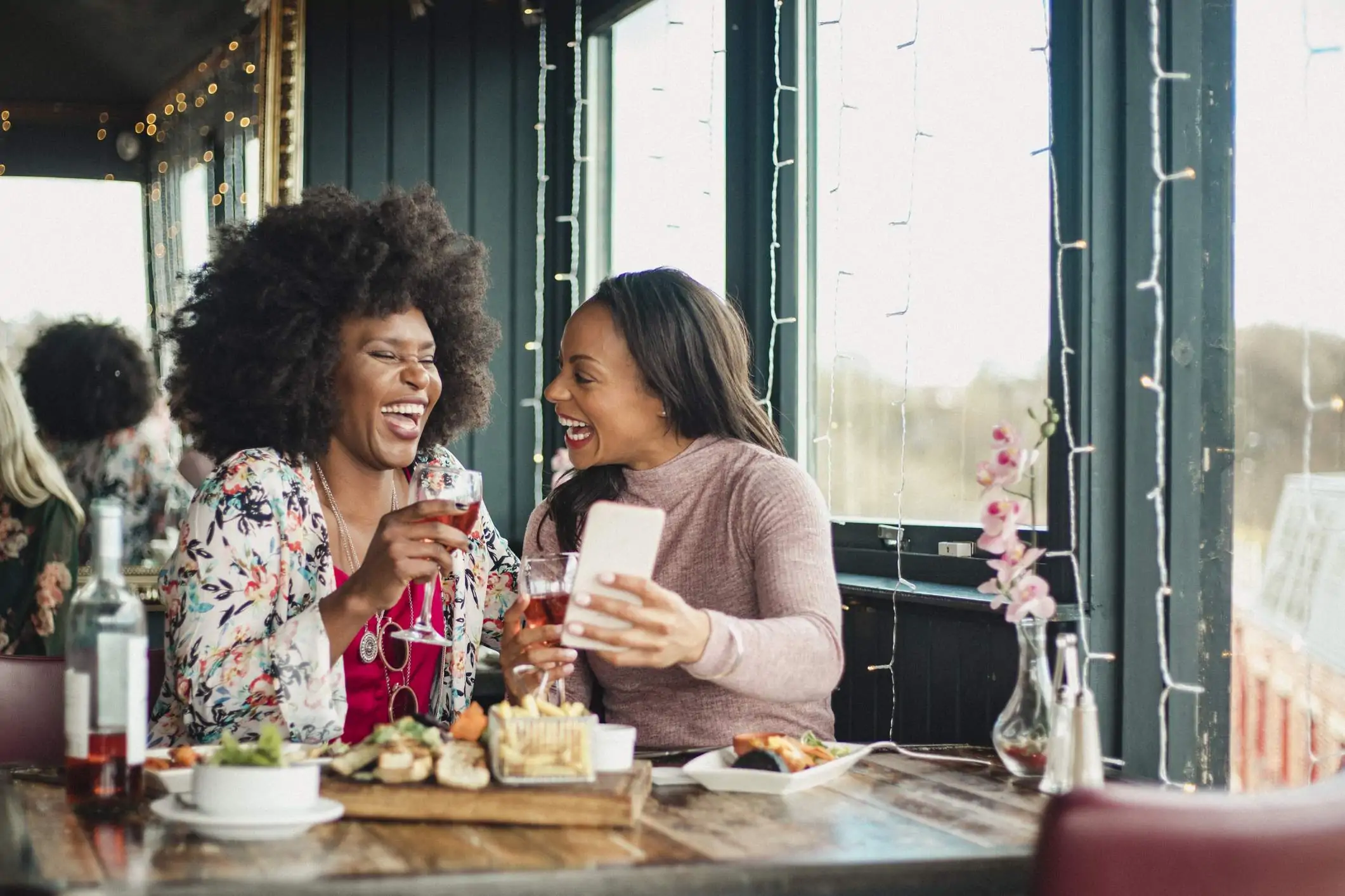 Image depicts two diners seated at a restaurant. They are sharing some plates and drinking wine. They are both laughing as one of them holds up their phone.