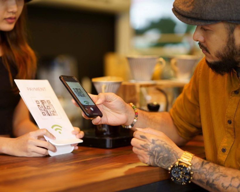 Image depicts two people at a counter. The host is holding up a QR code payment sign and the person who looks to be a customer is scanning the code with a mobile phone.