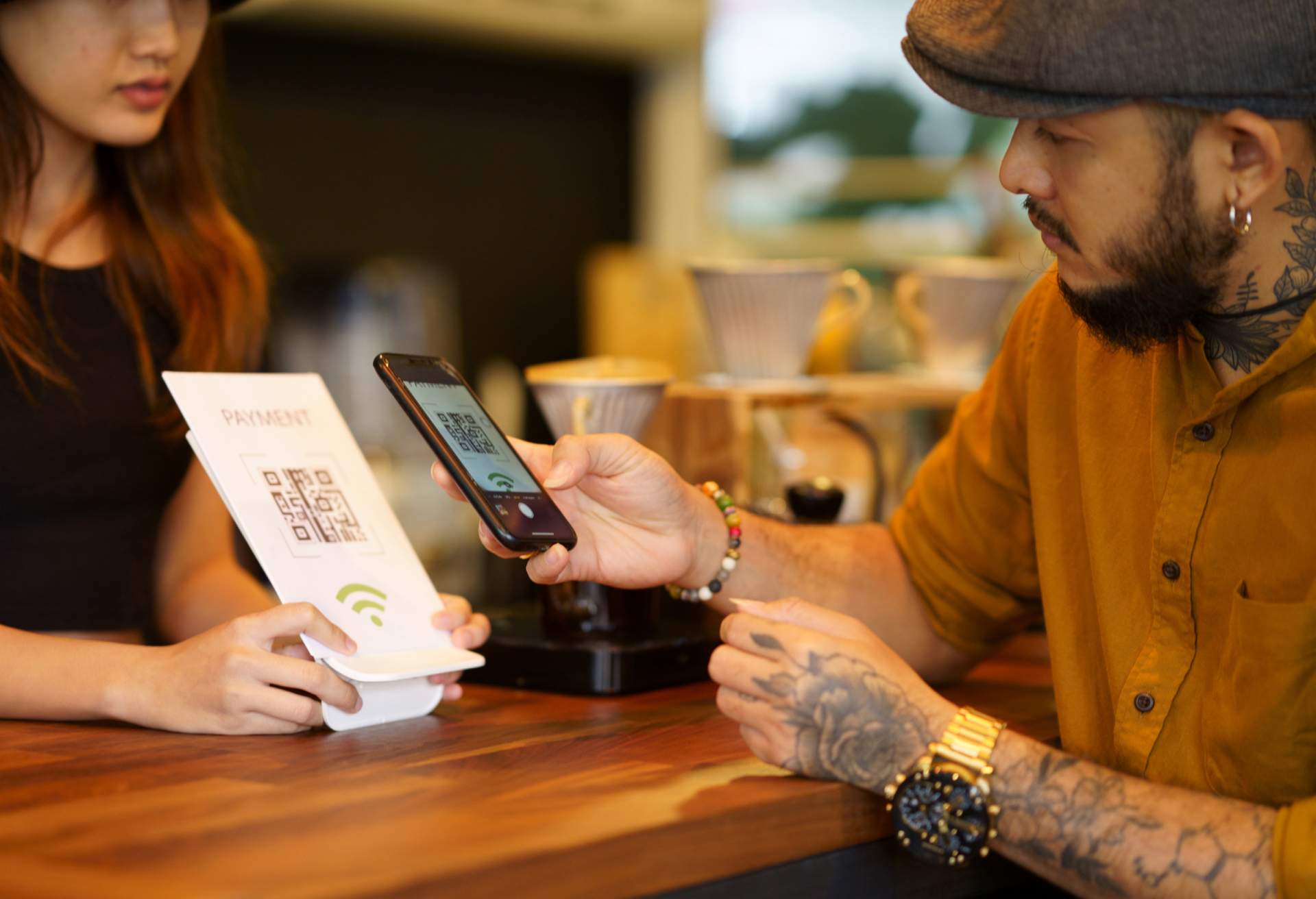 Image depicts two people at a counter. The host is holding up a QR code payment sign and the person who looks to be a customer is scanning the code with a mobile phone.