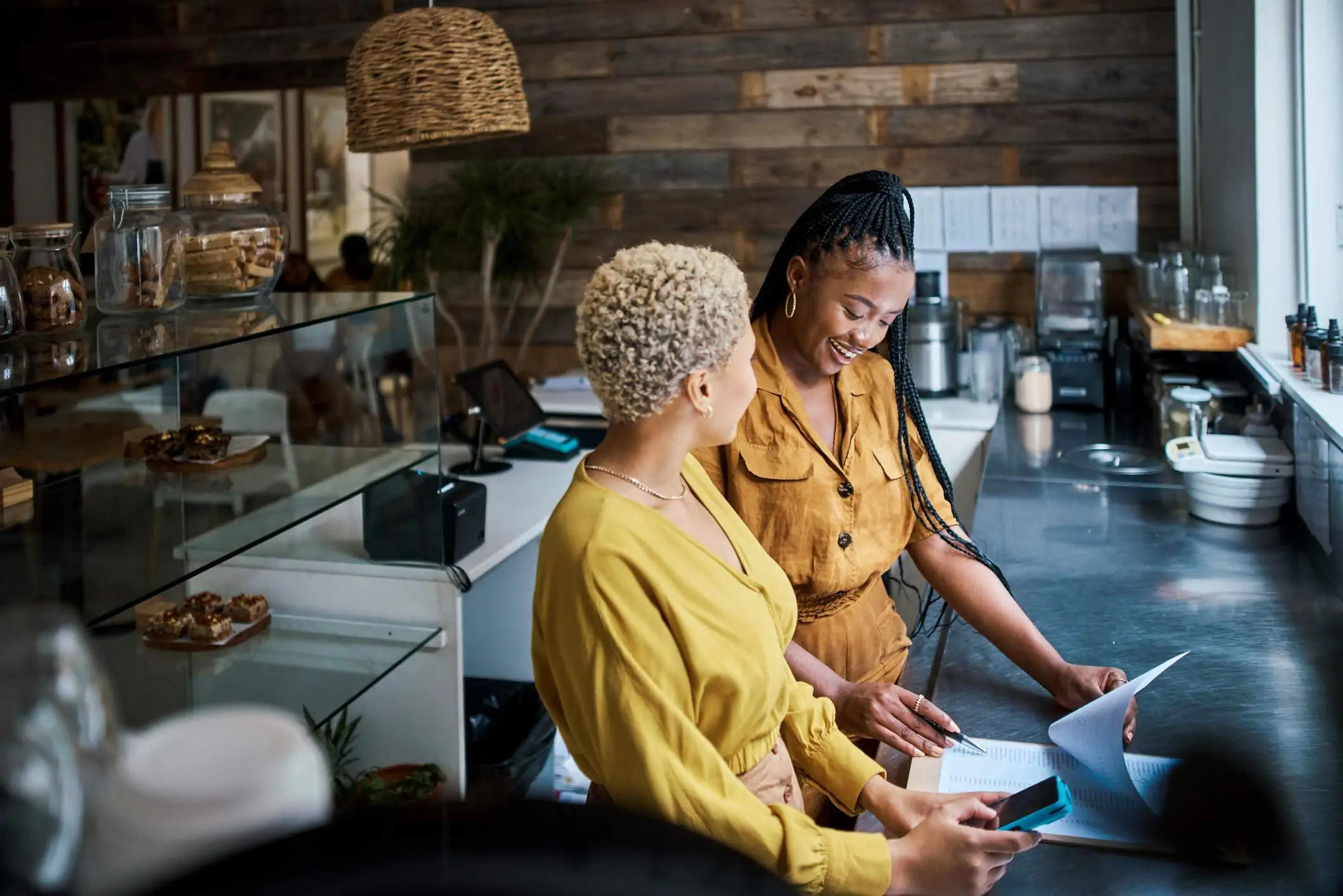 Image depicts two restaurant workers smiling. They are both wearing shades of yellow. One is smiling down at a clipboard as she is reviewing numbers on a paper. The other is holding a phone. There are pastries on display behind them.