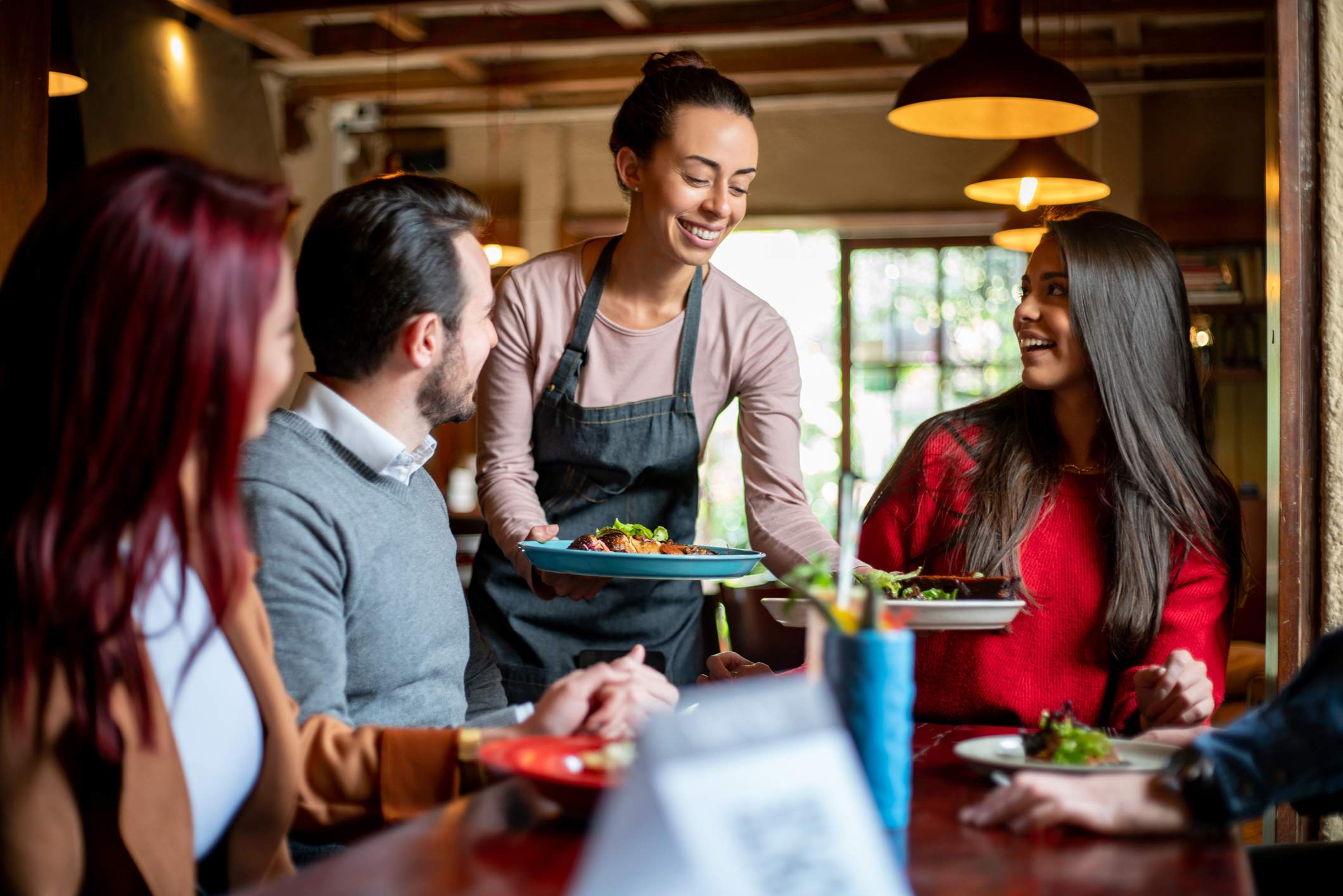 Image depicts a server bringing two plates of food to a table of people. The server is wearing a long sleeve shirt and a denim apron. The people at the table are of varying races and genders. They are all smiling. 