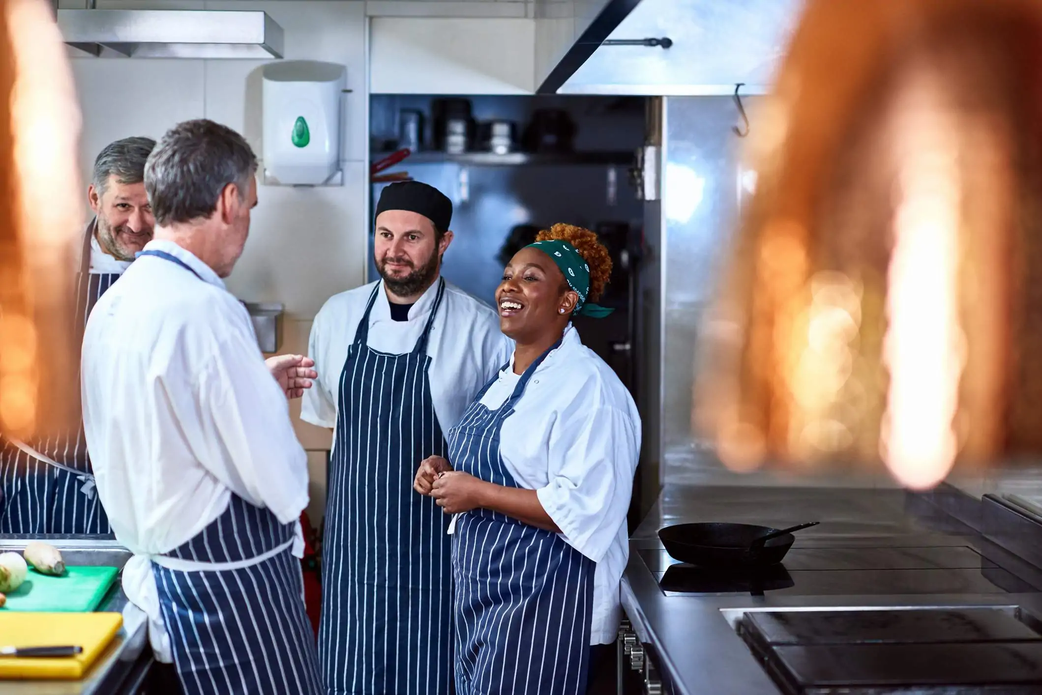 Image depicts four restaurant workers, all wearing white shirts and blue striped aprons, chatting in a kitchen.