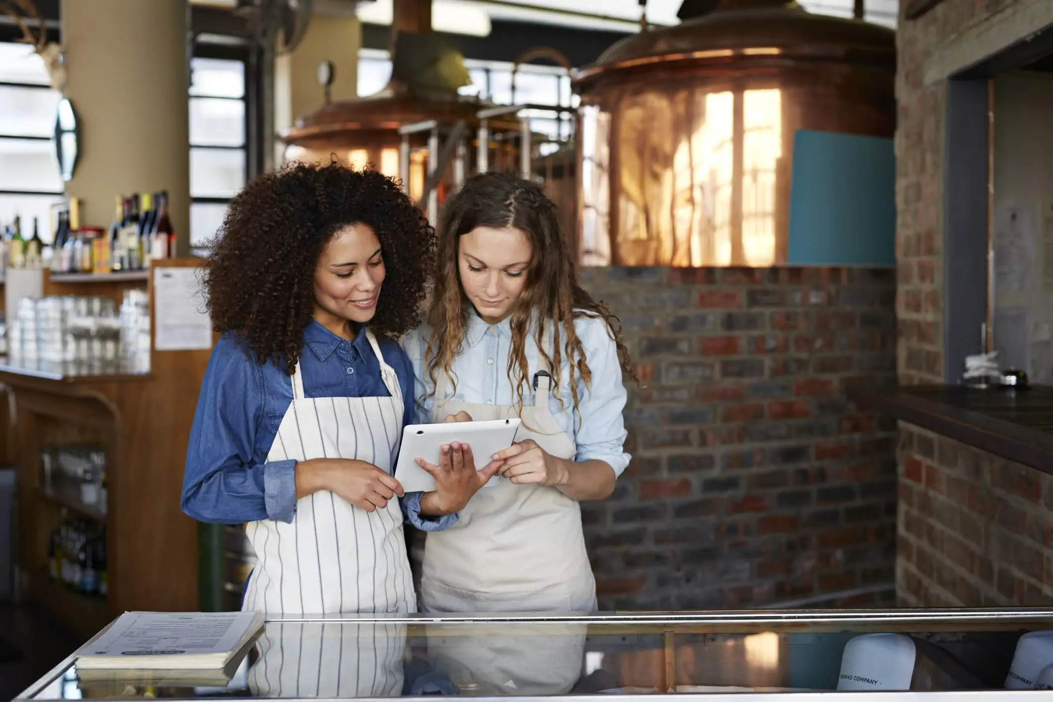 Image depicts two servers using a tablet. One is wearing a dark blue button down and a white striped apron. The other wears a light blue button down and a tan apron.