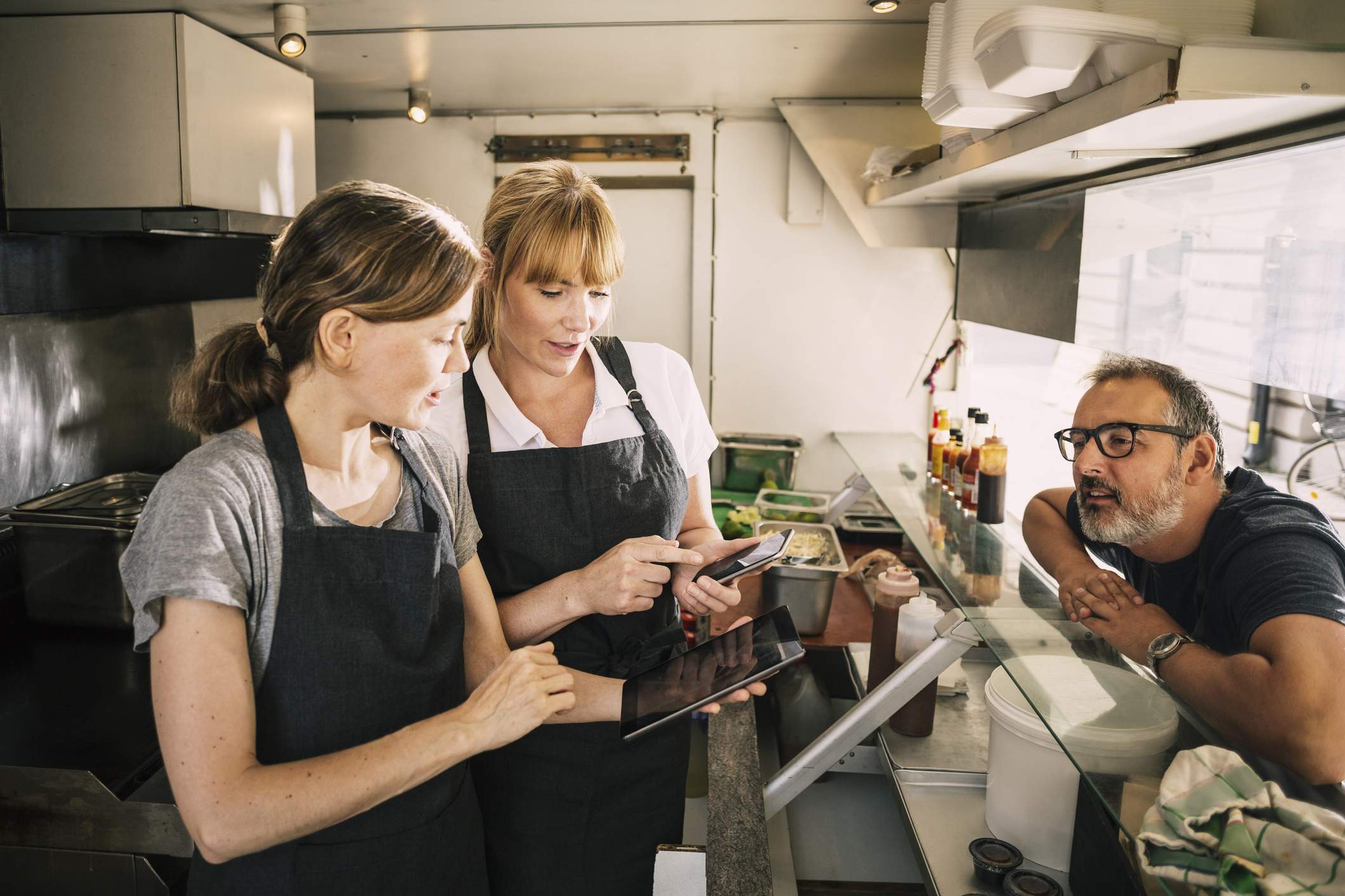 Image depicts two servers standing behind a counter. One, wearing a white polo shirt and black apron, is showing the other something on a phone. The other, dressed in a gray t-shirt and black apron, is holding a tablet. There is another worker wearing black rimmed glasses leaning over the counter. 