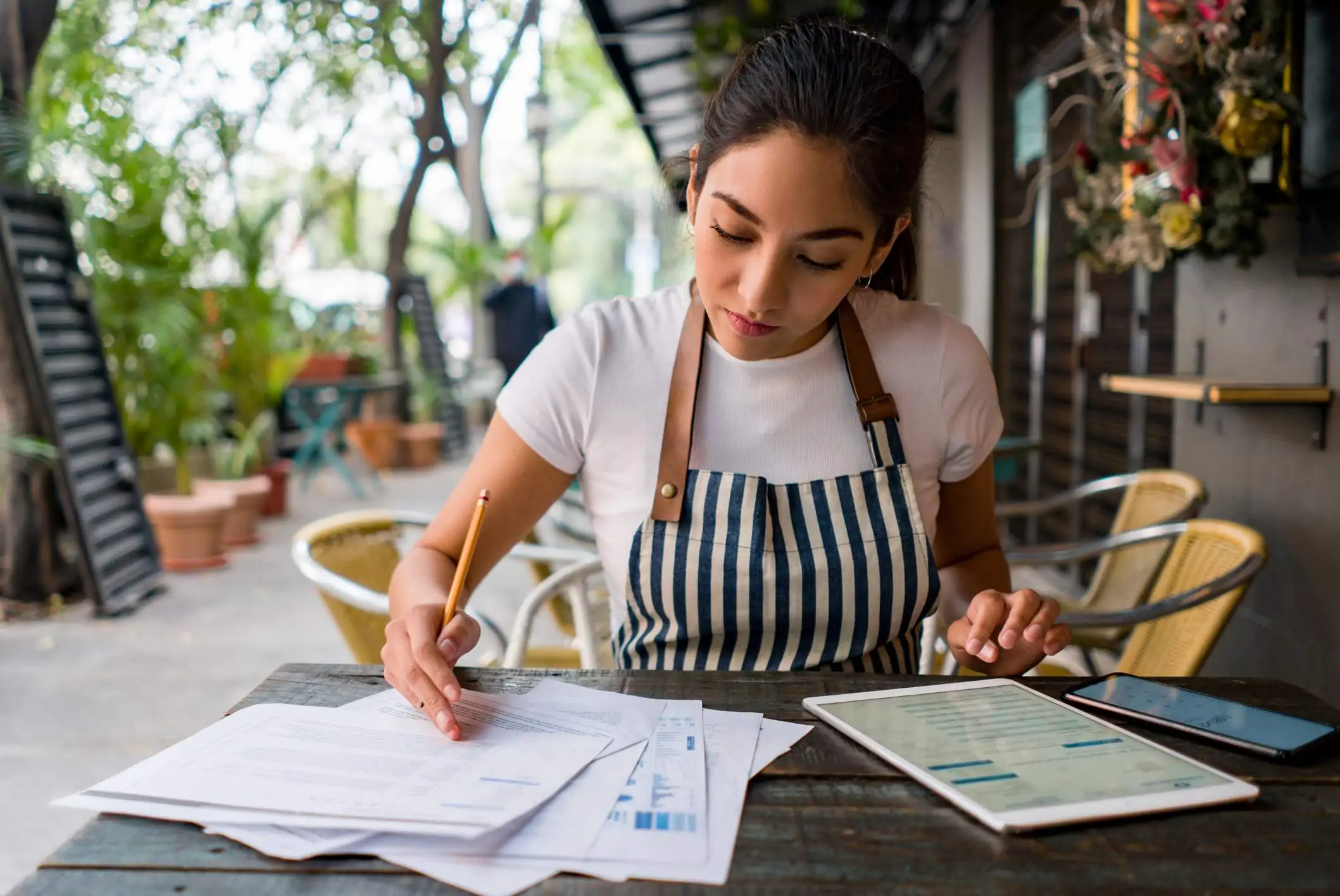 Image depicts a restaurant worker wearing a blue and white striped apron and a white shirt. They are sitting at a table outside. On the table is a tablet, a phone, and piles of paper. The worker is looking at some graphs on the paper and writing something with a pencil.