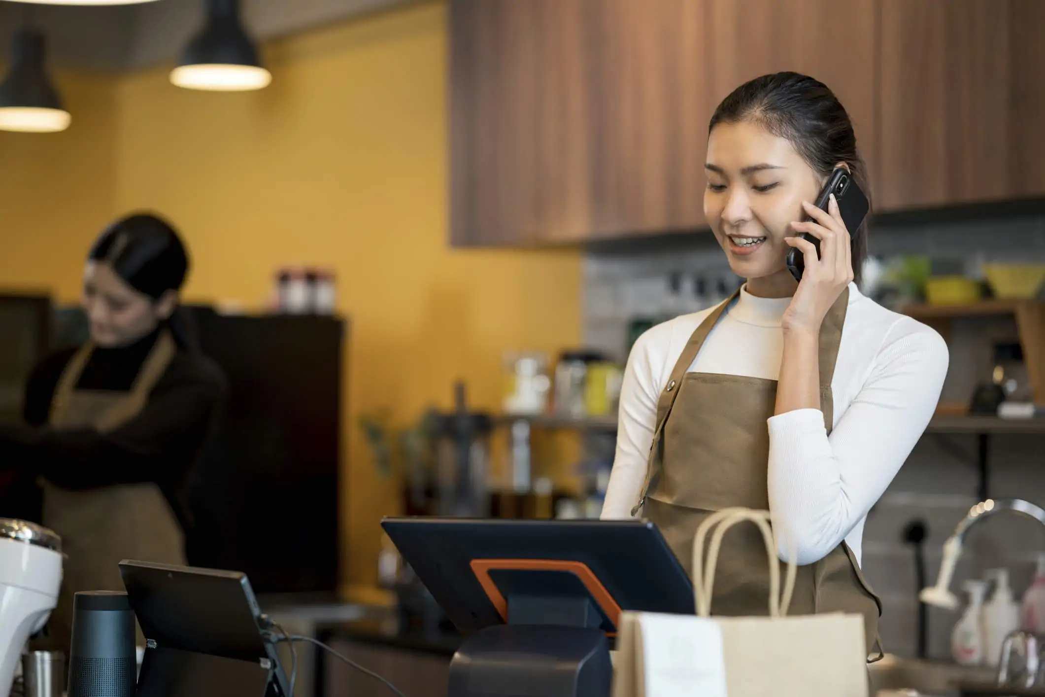 Image depicts a restaurant worker wearing a brown suede apron and long-sleeved white shirt. They are talking on a mobile phone and are using a mounted tablet as a point of sale (POS) system.