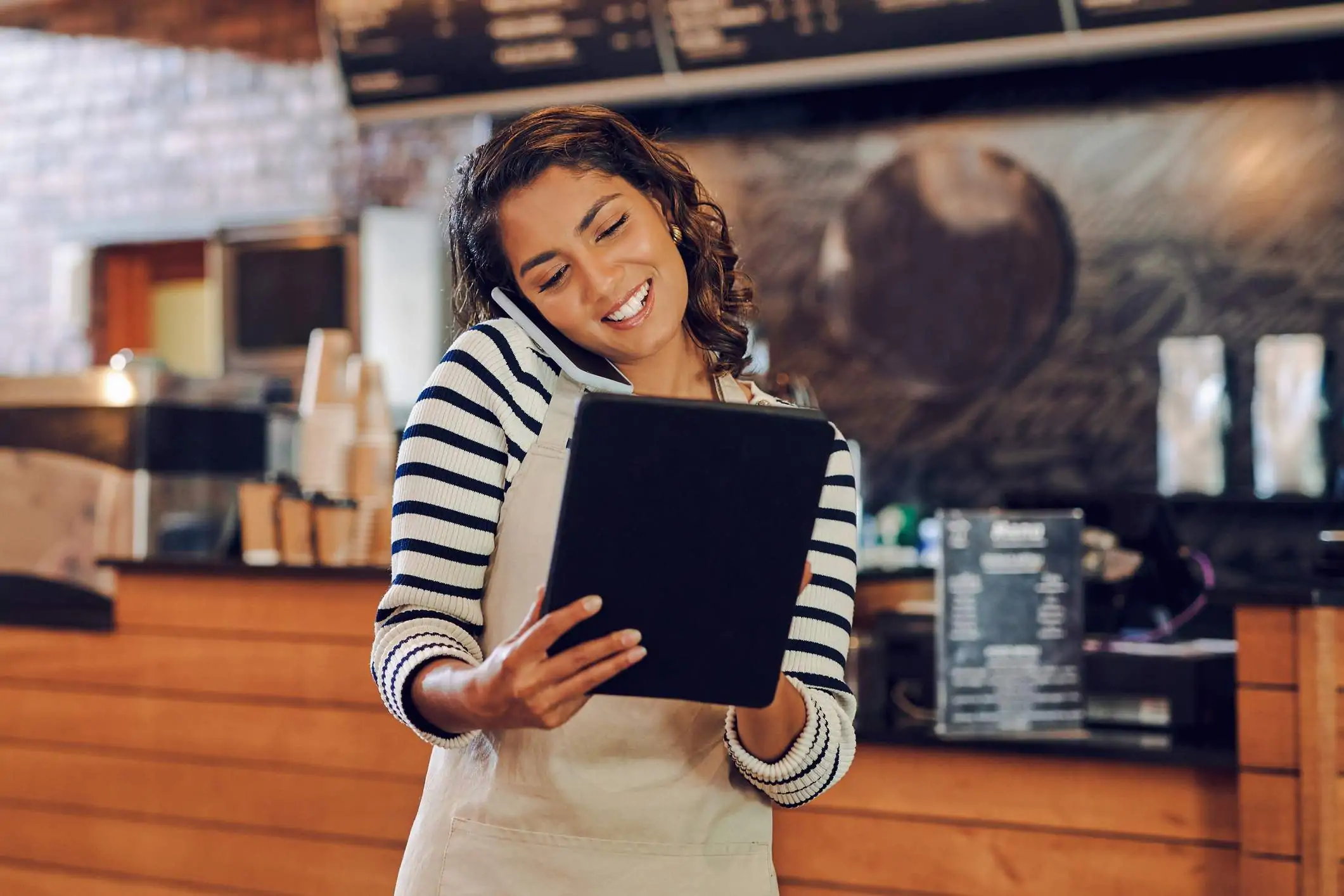 Image depicts a restaurant worker wearing a beige apron and a blue and white striped shirt. They are talking on the phone and are using a tablet.