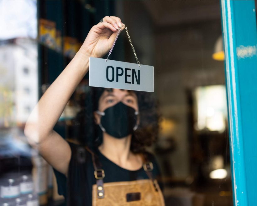 Restaurant employee in black apron behind a glass door flips a sign to read “open”