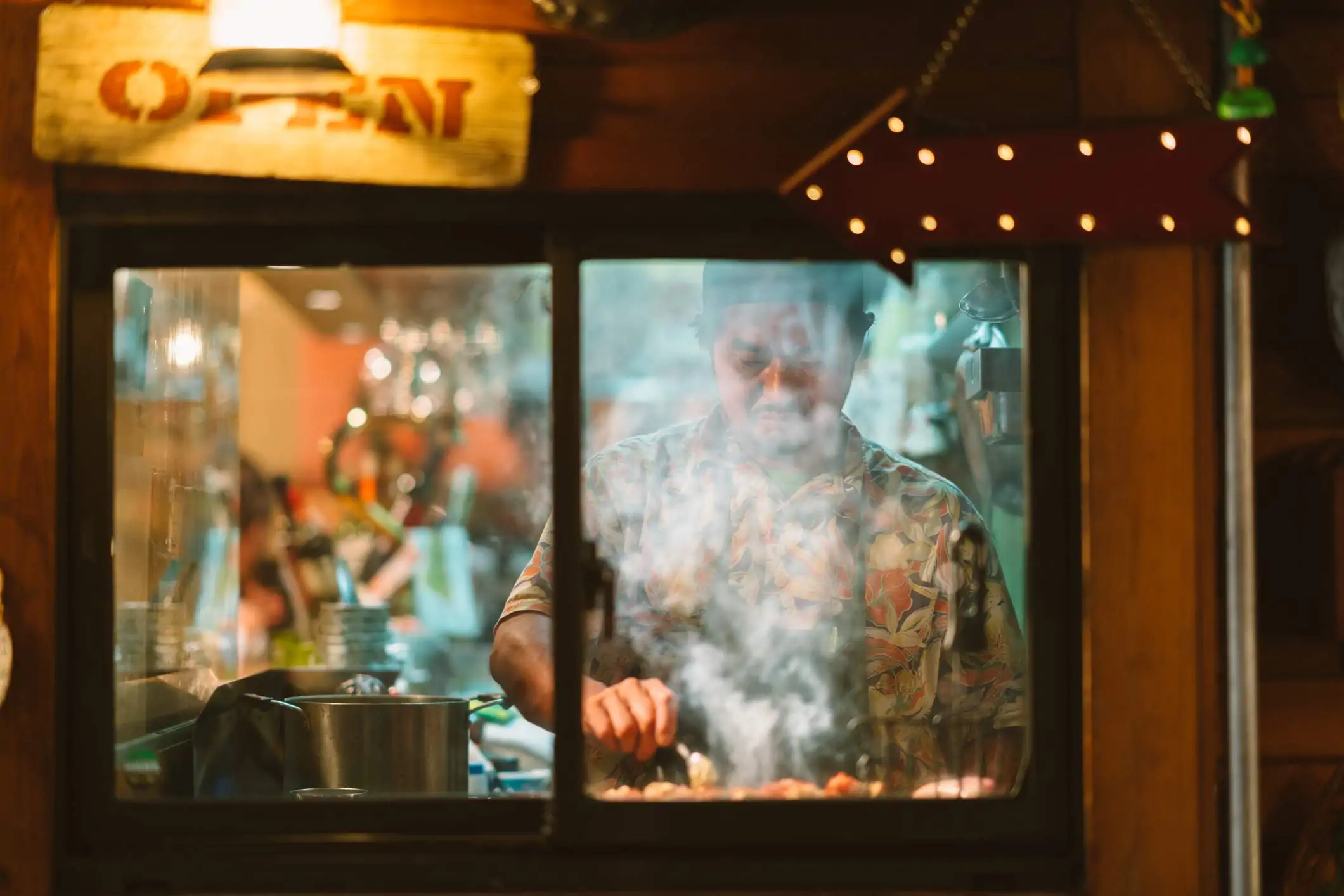 Image depicts a chef wearing a floral shirt and an apron cooking behind a window. There is steam coming up from the meal they are preparing. There is an open sign hanging above the window.