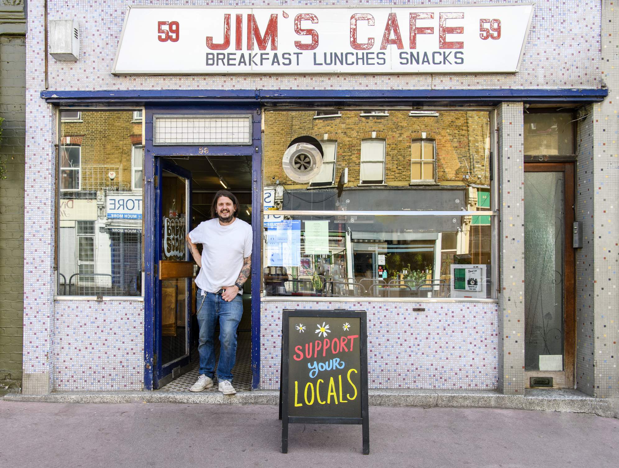 Image depicts a person wearing a white shirt and jeans standing in the doorway. A sign over the establishment reads “Jim’s Cafe, breakfast, lunch, snacks.” There are two large windows and the front is tiled with mosaic. A kick sign out front reads “support your locals.”