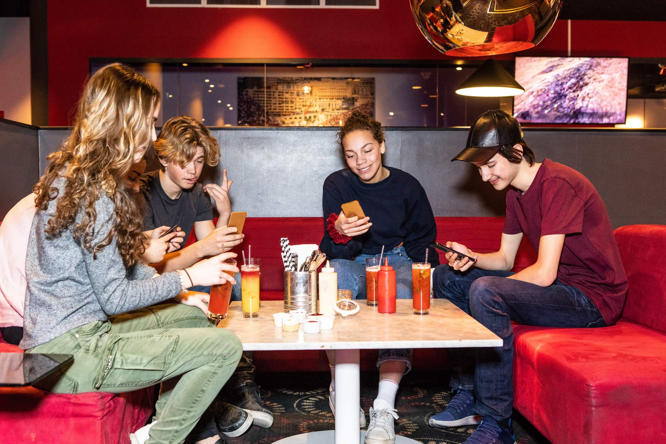 Image depicts a group of young people sitting in a booth. Many are looking at their phones and they are sipping mocktails.